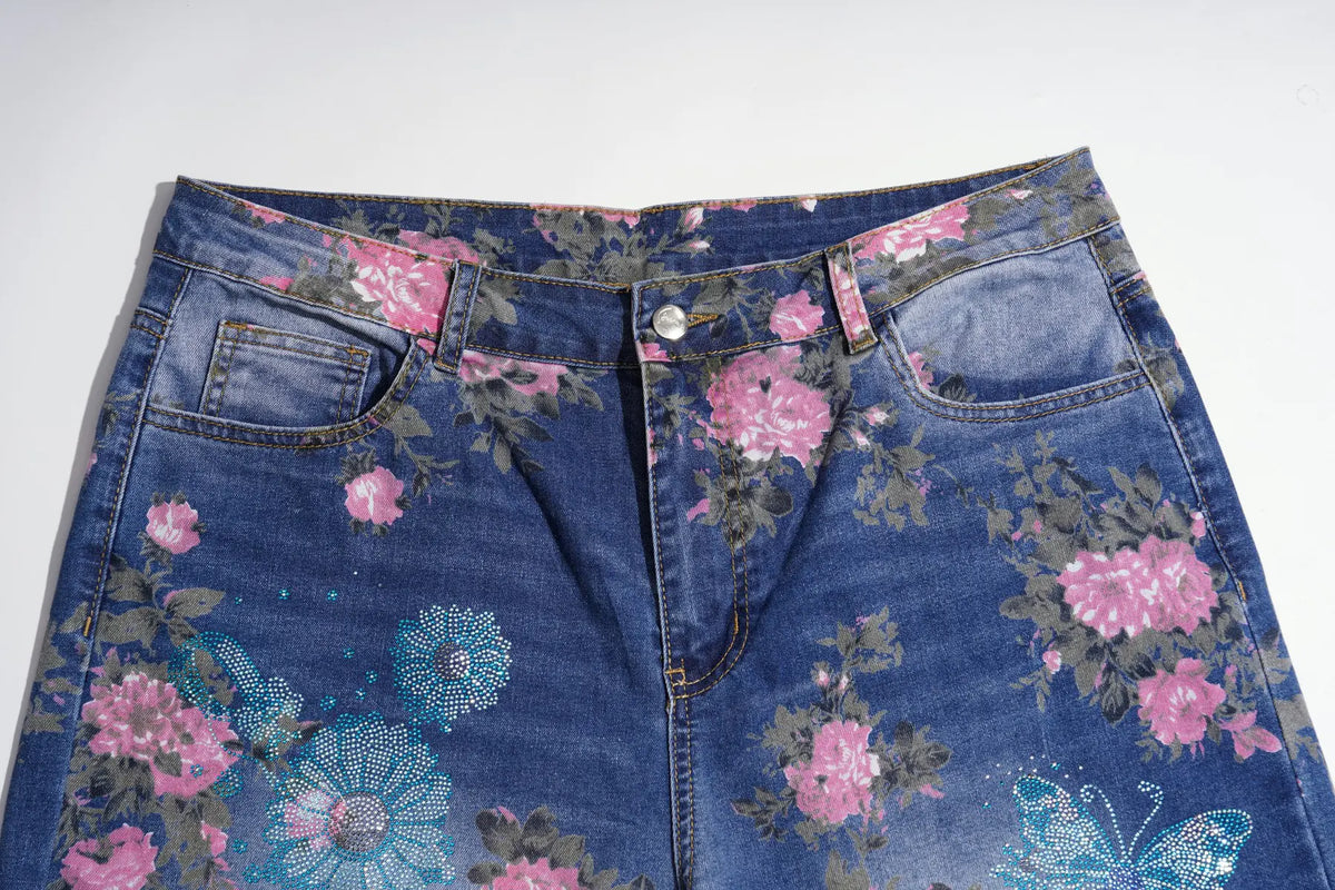 Close-up of a pair of blue denim jeans with a pink, teal, and gray floral print, laid flat against a white background. TCH Blue Floral Butterfly Rhinestone Wide-Leg Jeans