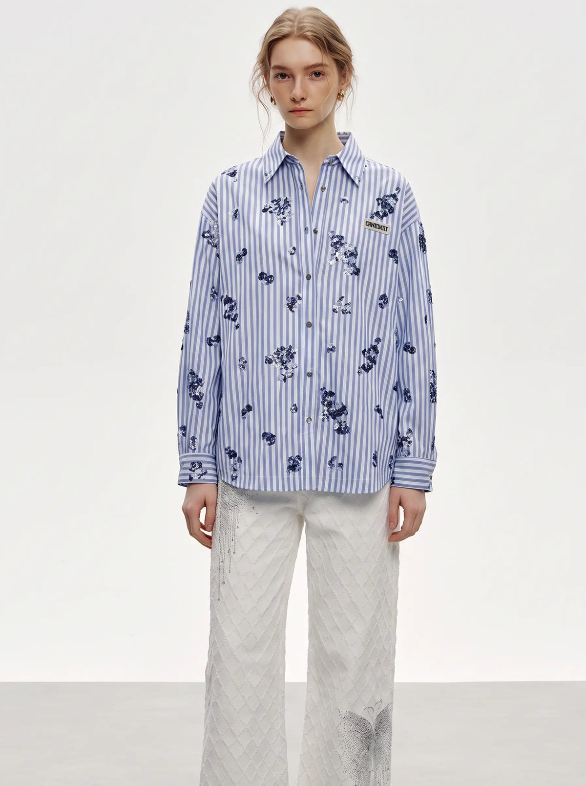 A model stands against a light gray studio background wearing a blue and white vertically striped button-up shirt with a dark blue floral print, paired with white textured wide-leg pants. TCH Blue Floral Embroidered Pinstripe Shirt
