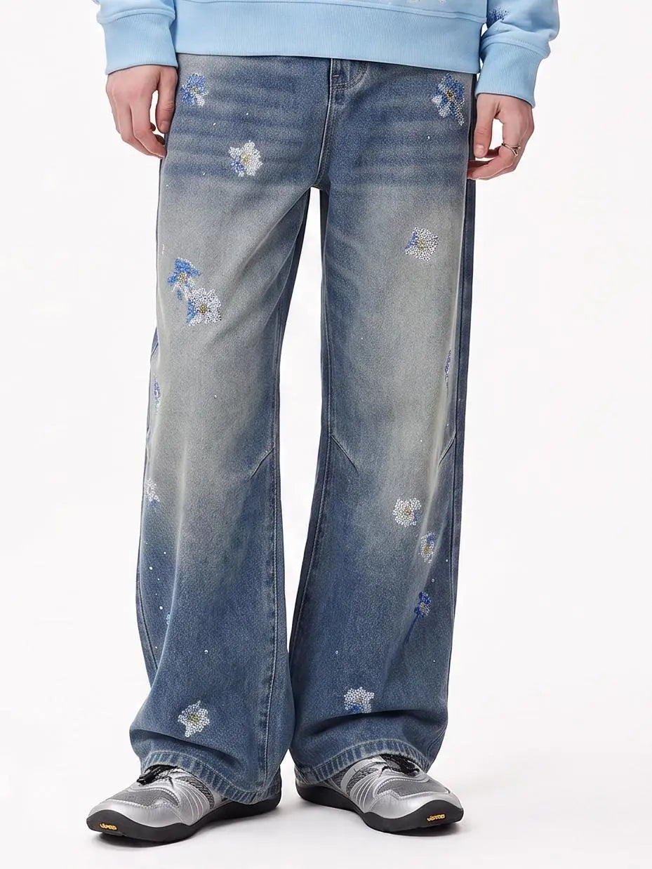 A person wearing wide-leg blue denim jeans with distressed fading and scattered embroidered white and blue floral details, paired with a light blue sweatshirt and gray sneakers, against a plain white studio background. TCH Blue Floral Rhinestone Wide-Leg Jeans