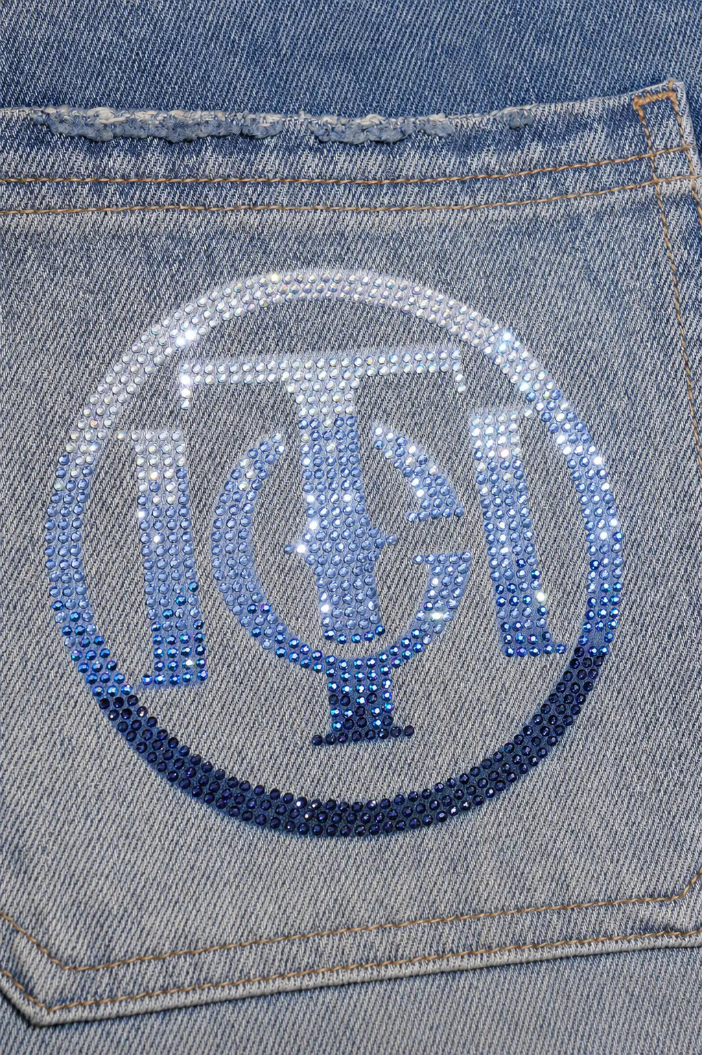 A close-up of a light blue denim pocket with a circular monogram patch made of blue and silver rhinestones on the front. TCH Blue Floral Rhinestone Wide-Leg Jeans