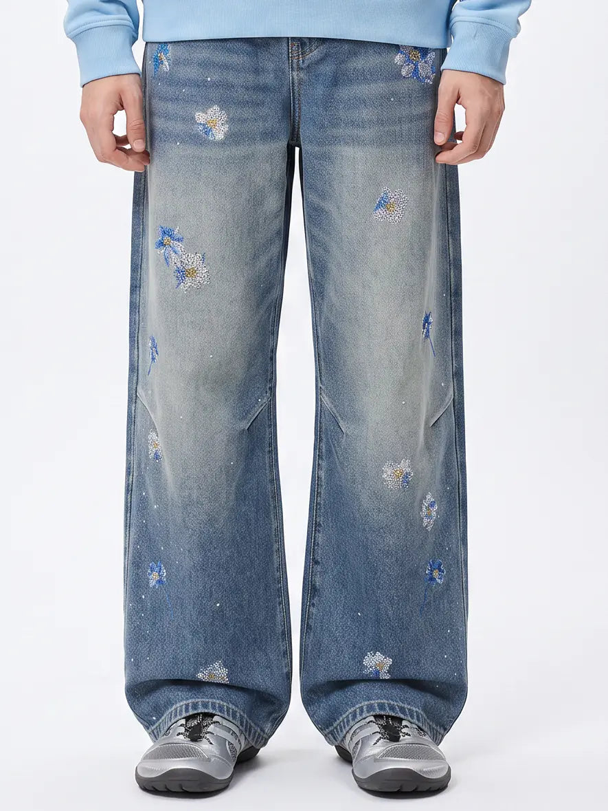 A person wearing wide-leg blue denim jeans with scattered embroidered white and blue floral details, paired with a light blue sweatshirt and gray sneakers, shown from the waist down on a white studio background. TCH Blue Floral Rhinestone Wide-Leg Jeans