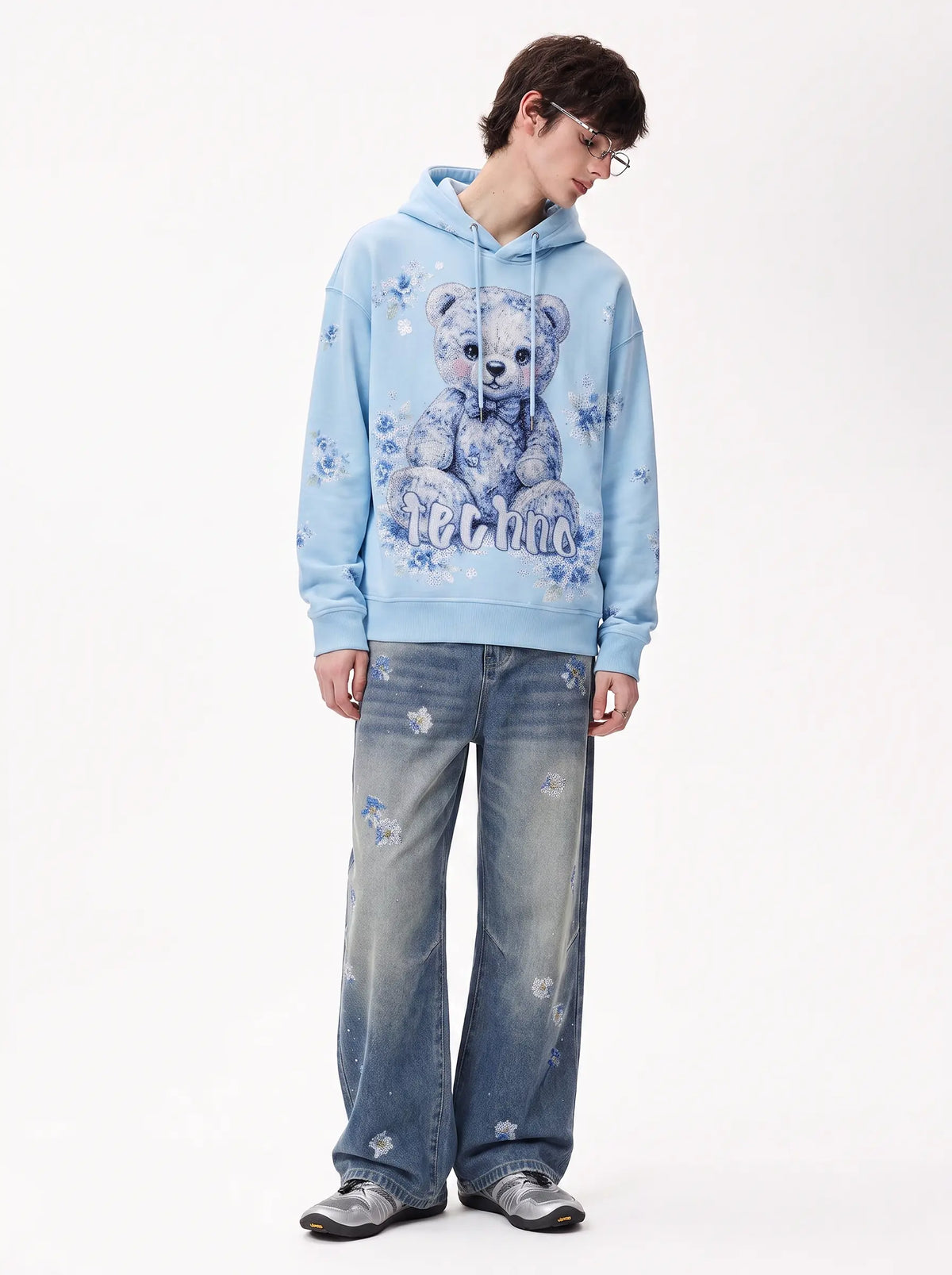 A model wearing a light blue hoodie with a teddy bear graphic and distressed blue jeans, standing against a plain white studio background. TCH Blue Floral Rhinestone Wide-Leg Jeans