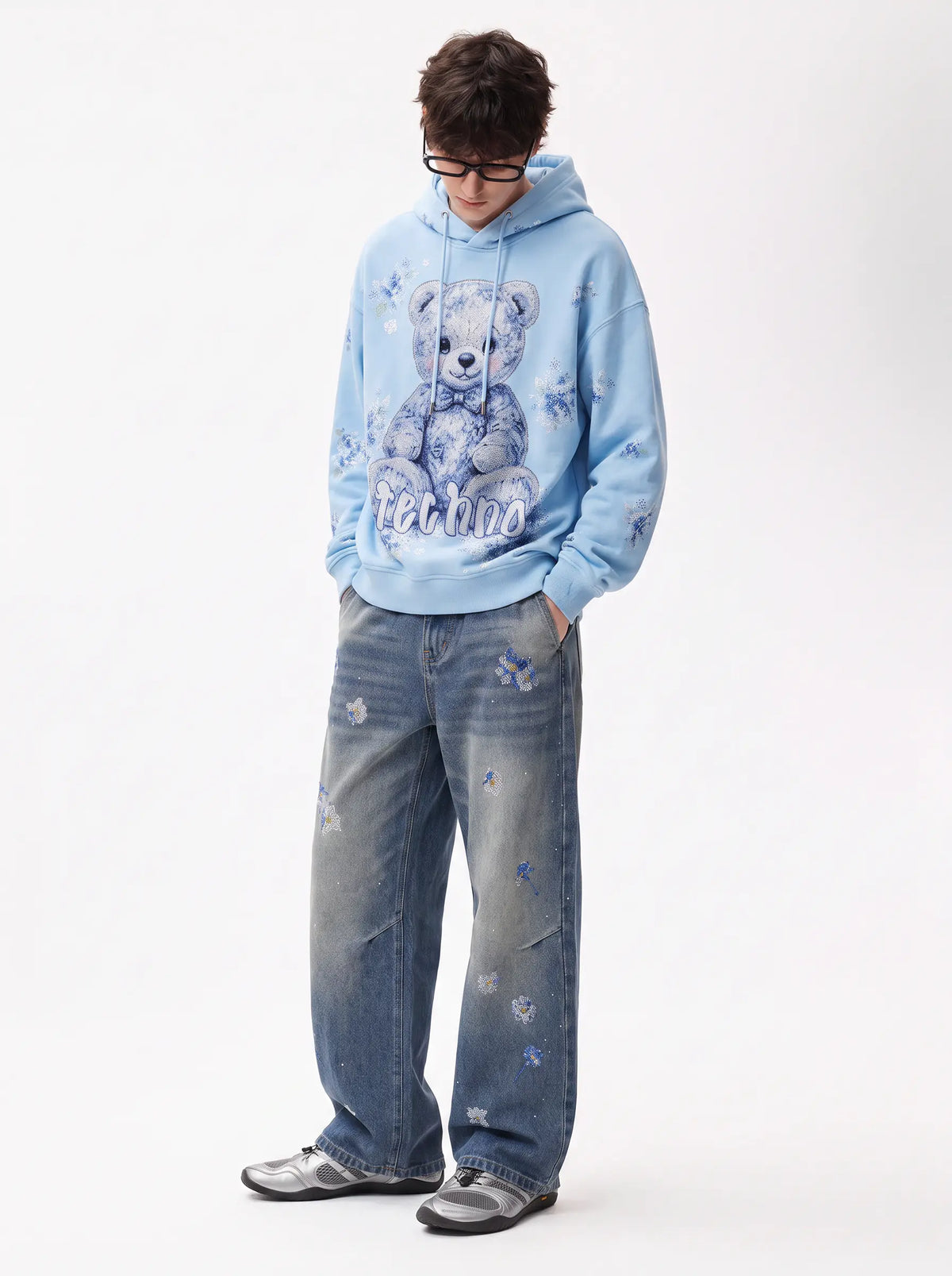 A model wearing a light blue hoodie with a teddy bear graphic and distressed blue jeans, standing against a plain white studio background. TCH Blue Floral Rhinestone Wide-Leg Jeans