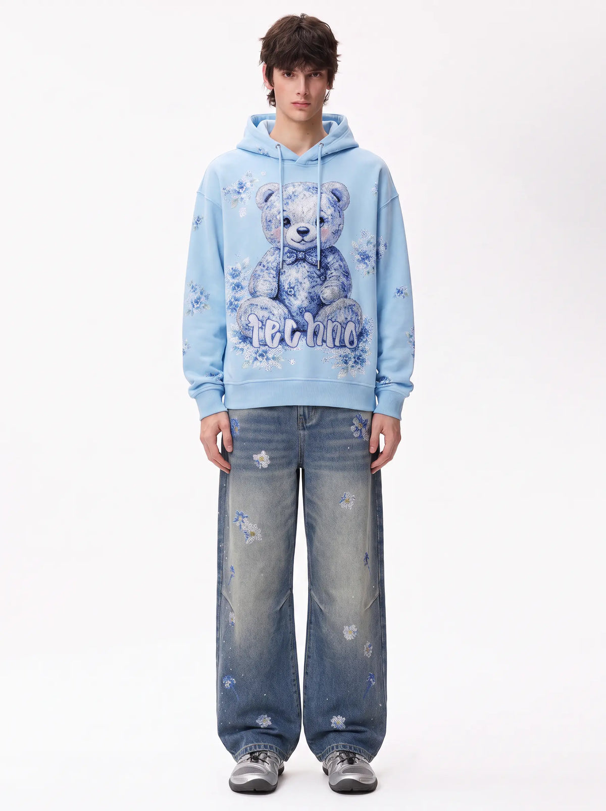 A model stands against a white studio background wearing a light blue hoodie with a large blue teddy bear graphic and the word 'petite' on the front, paired with wide-leg distressed blue jeans and gray sneakers. TCH Blue Floral Rhinestone Wide-Leg Jeans