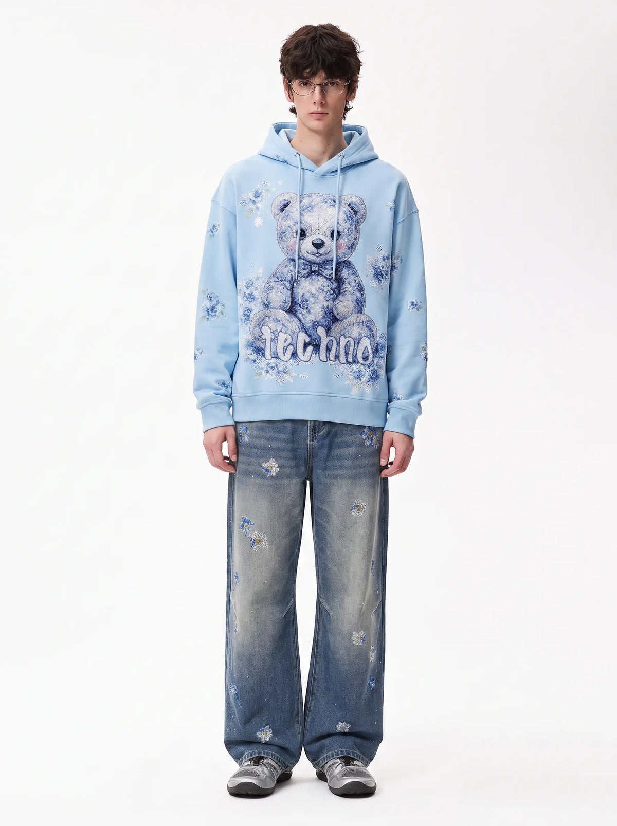 A model stands against a white studio background wearing a light blue hoodie with a large teddy bear graphic and the word 'Missoni' printed on the front, paired with distressed blue jeans and gray sneakers. TCH Blue Floral Rhinestone Wide-Leg Jeans
