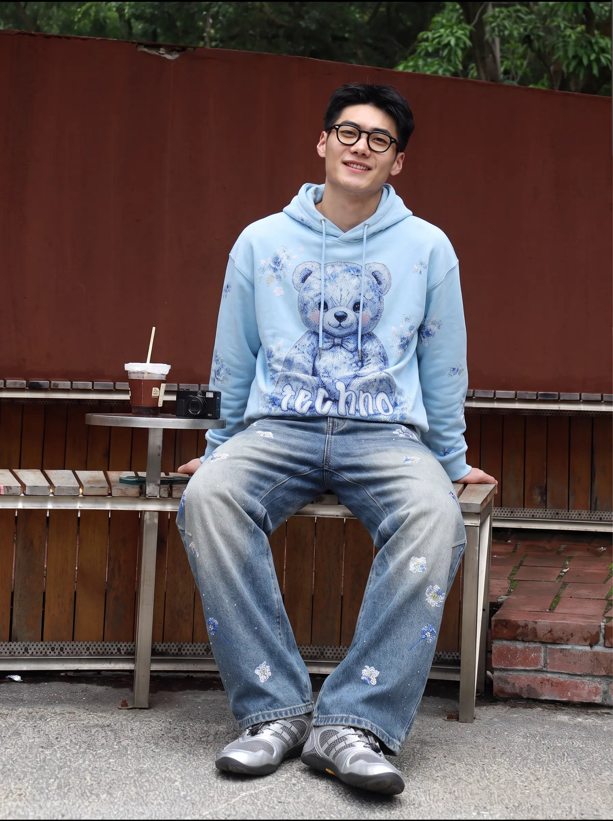A man wearing a light blue hoodie with a teddy bear graphic and matching distressed blue jeans, sitting on a wooden bench outdoors. TCH Blue Floral Rhinestone Wide-Leg Jeans