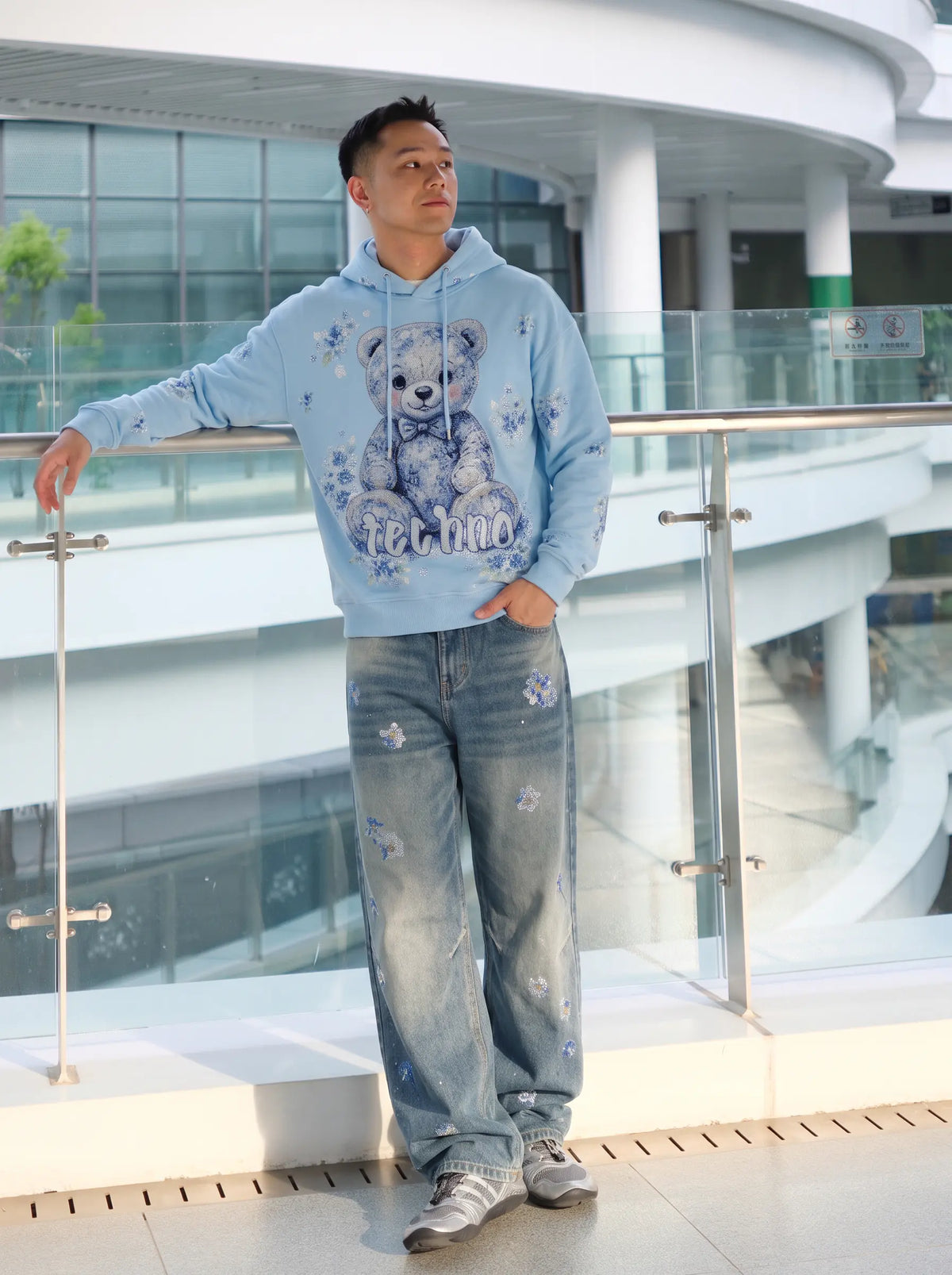 A man wearing a light blue hoodie with a teddy bear graphic and matching light blue jeans, standing on an outdoor balcony with a glass railing. TCH Blue Floral Rhinestone Wide-Leg Jeans