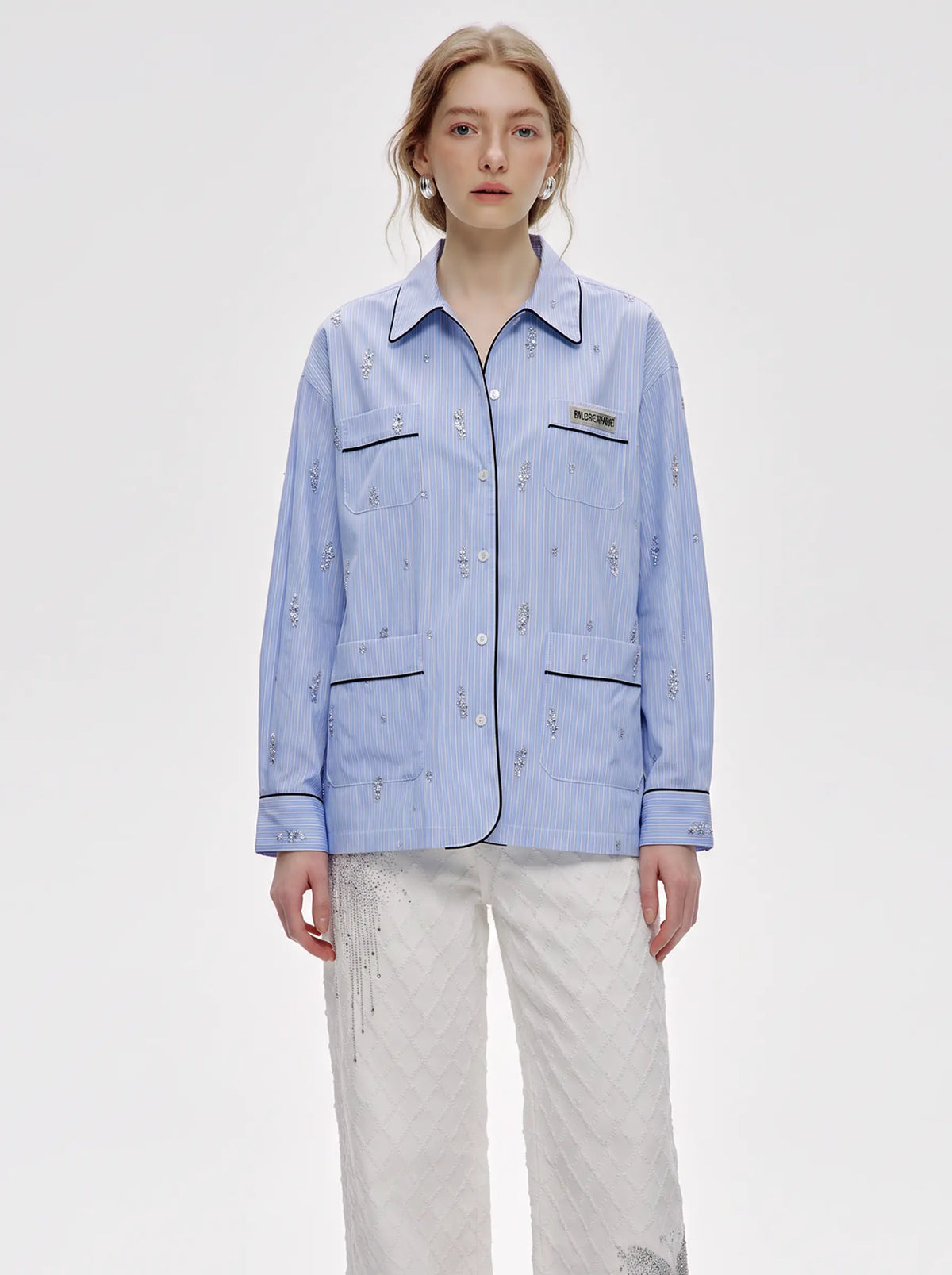 A model wearing a light blue striped button-up shirt with embroidered details and white pants, photographed against a plain light gray studio background. TCH Blue Pinstripe Embroidered Pocket Shirt