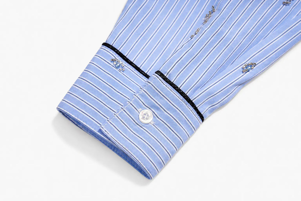 Close-up of a light blue and white striped dress shirt cuff with black piping, a white button, and embroidered details on a white background. TCH Blue Pinstripe Embroidered Pocket Shirt