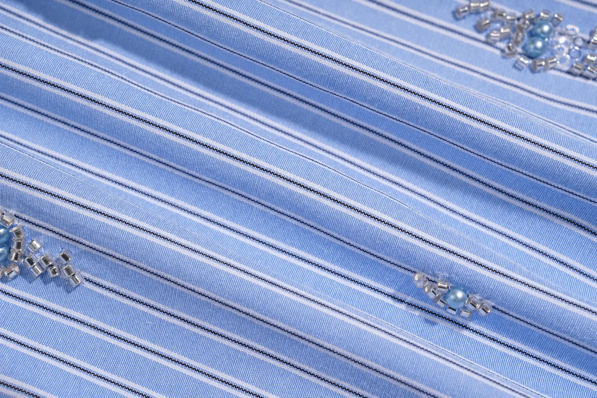 A close-up of light blue pleated fabric with thin dark pinstripes and small silver embellishments with blue pearls. TCH Blue Pinstripe Embroidered Pocket Shirt