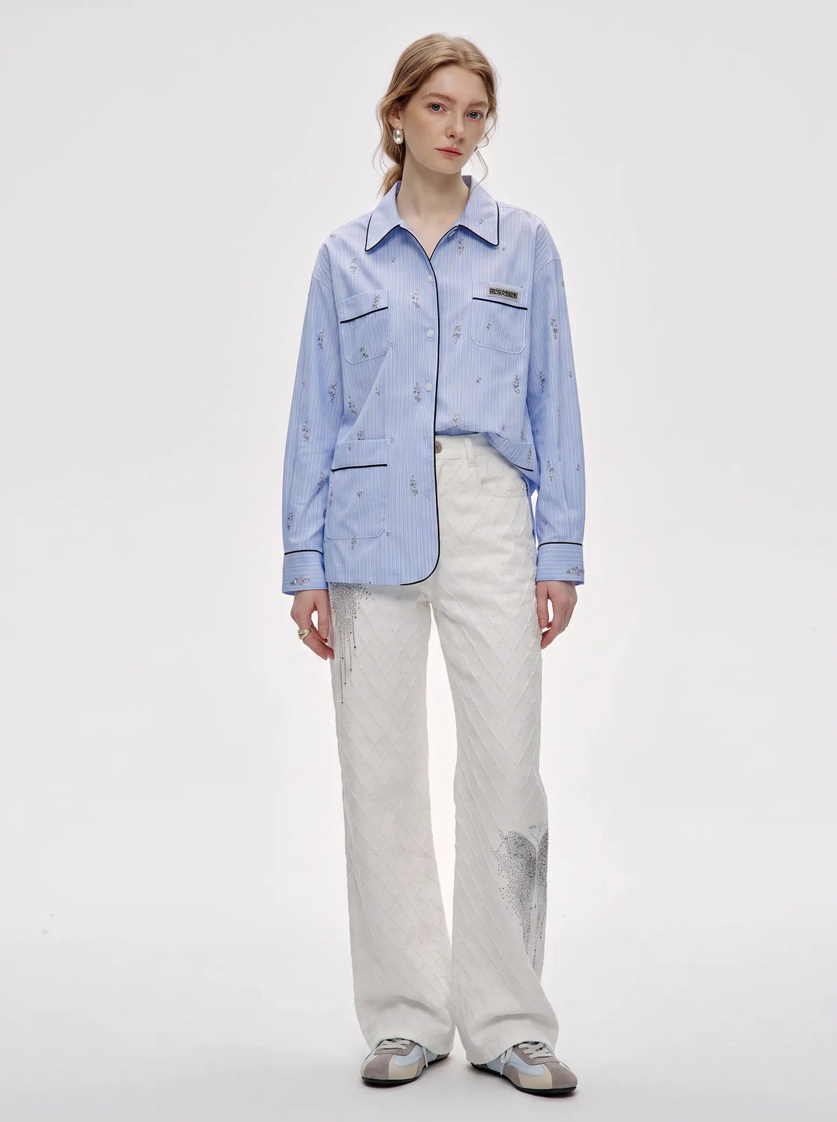 A model stands against a light gray studio background wearing a light blue button-up shirt with subtle tonal embroidery, paired with off-white wide-leg pants and gray sneakers. TCH Blue Pinstripe Embroidered Pocket Shirt