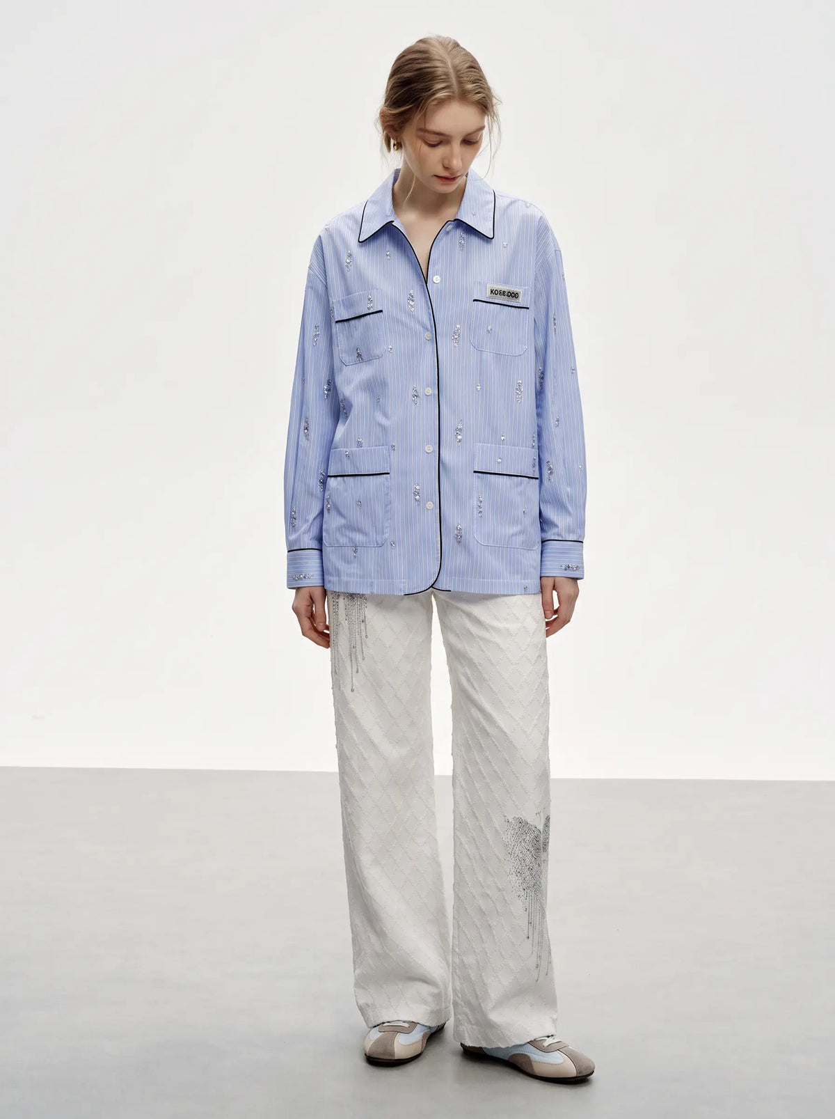 A model stands in a studio wearing a light blue embroidered long-sleeve button-front shirt jacket with flap pockets, paired with off-white wide-leg trousers and metallic flats. TCH Blue Pinstripe Embroidered Pocket Shirt