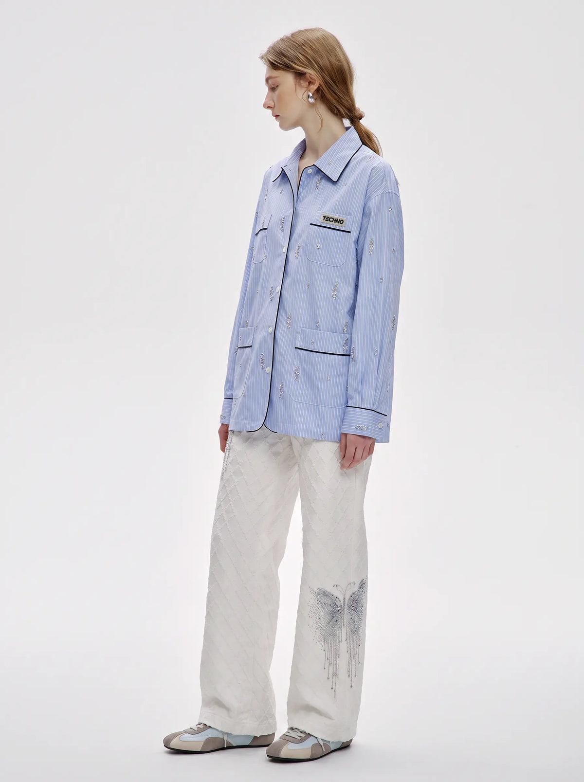 A woman stands in profile wearing a light blue button-up shirt jacket with embroidered details, paired with off-white wide-leg pants and gray slip-on shoes, against a plain light gray studio background. TCH Blue Pinstripe Embroidered Pocket Shirt