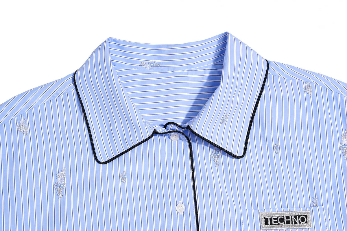 Close-up of a light blue and white striped button-down shirt with black piping trim on the collar and placket, featuring small embroidered motifs and a visible TECHNO brand patch on the chest. TCH Blue Pinstripe Embroidered Pocket Shirt