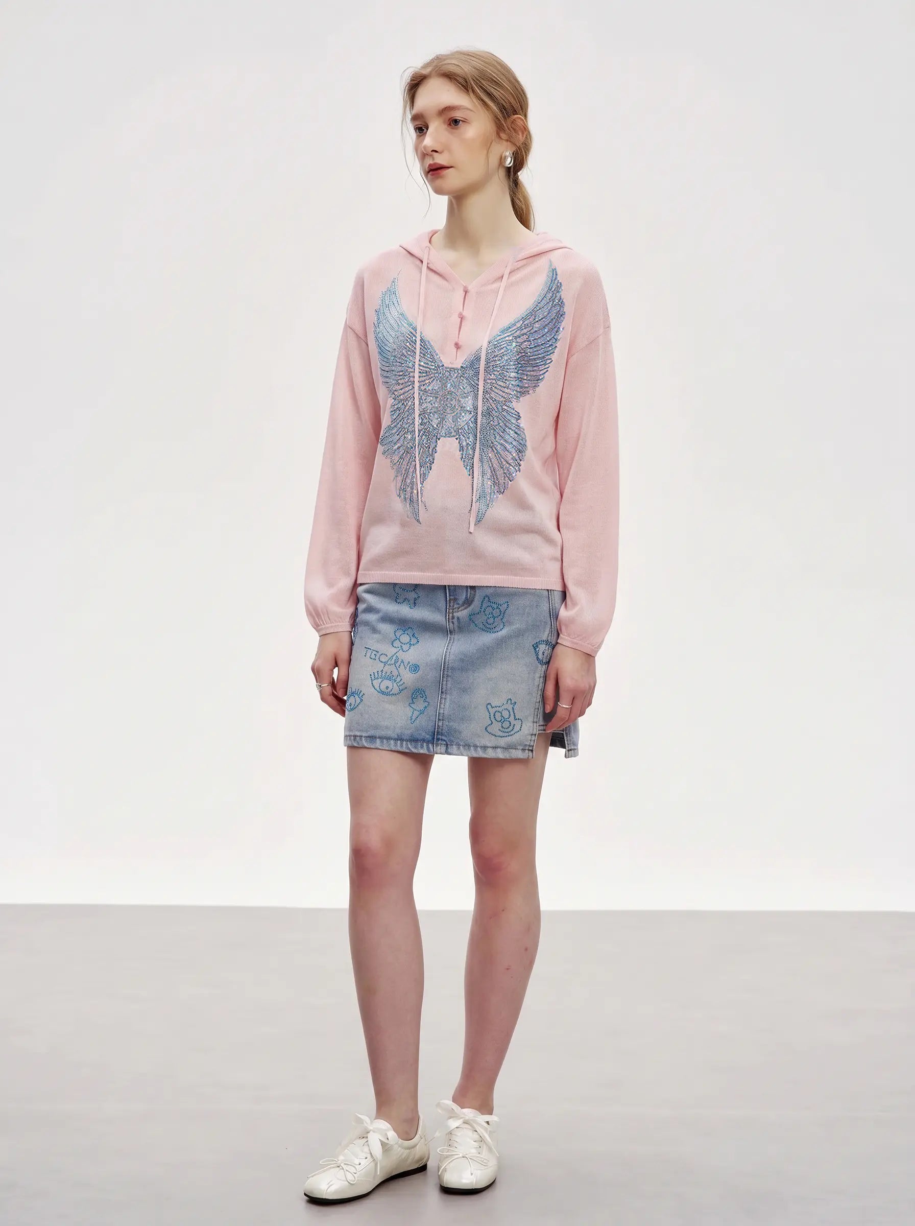 A model stands in a studio wearing a light pink hoodie with a large blue wing graphic on the front, paired with a light blue denim mini skirt and white sneakers. TCH Blue Rhinestone Angel Wings Cropped Hoodie