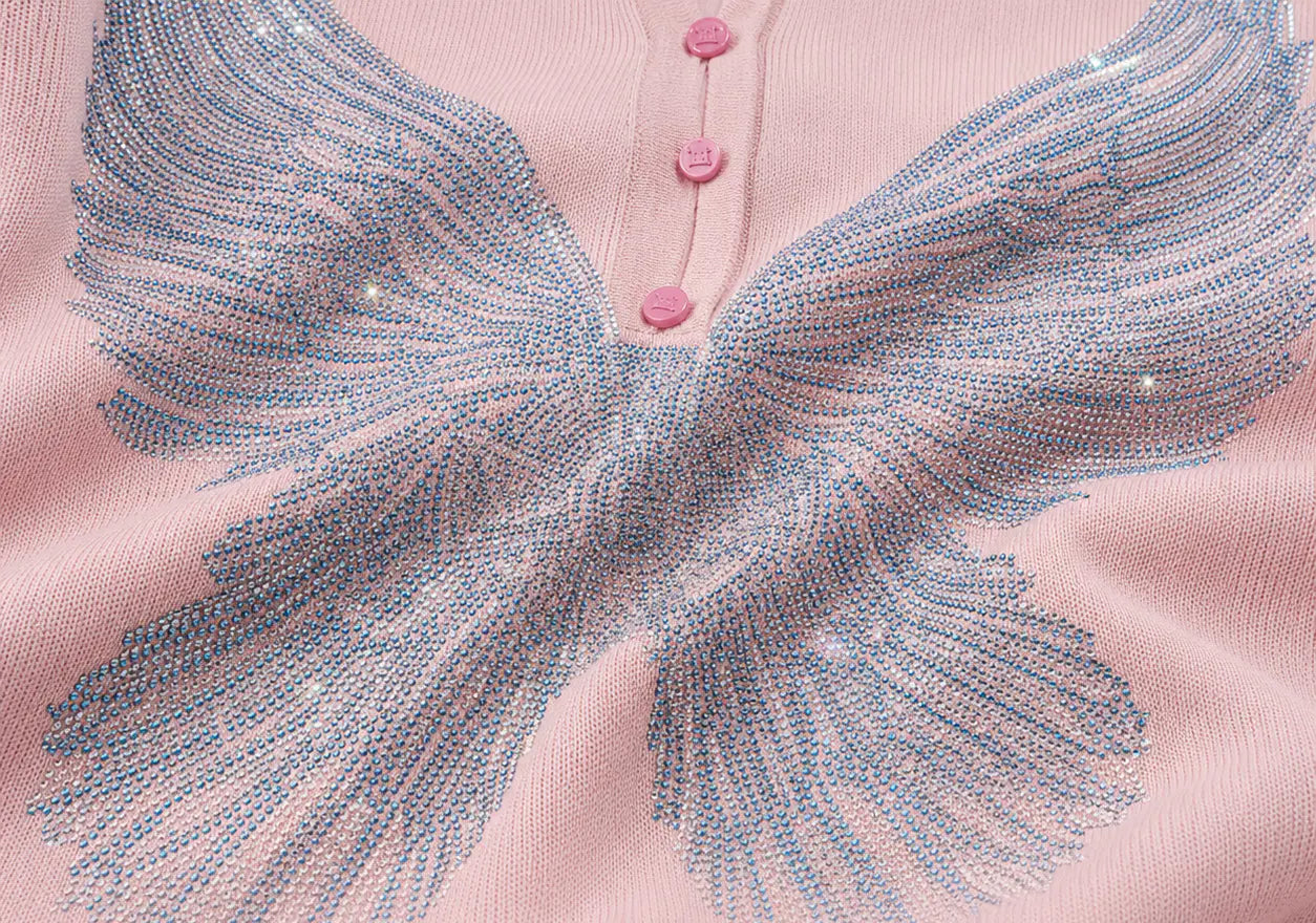 A close-up of a light pink knit top featuring a large embroidered wing design in light blue and silver thread, with three pink buttons at the neckline. TCH Blue Rhinestone Angel Wings Cropped Hoodie