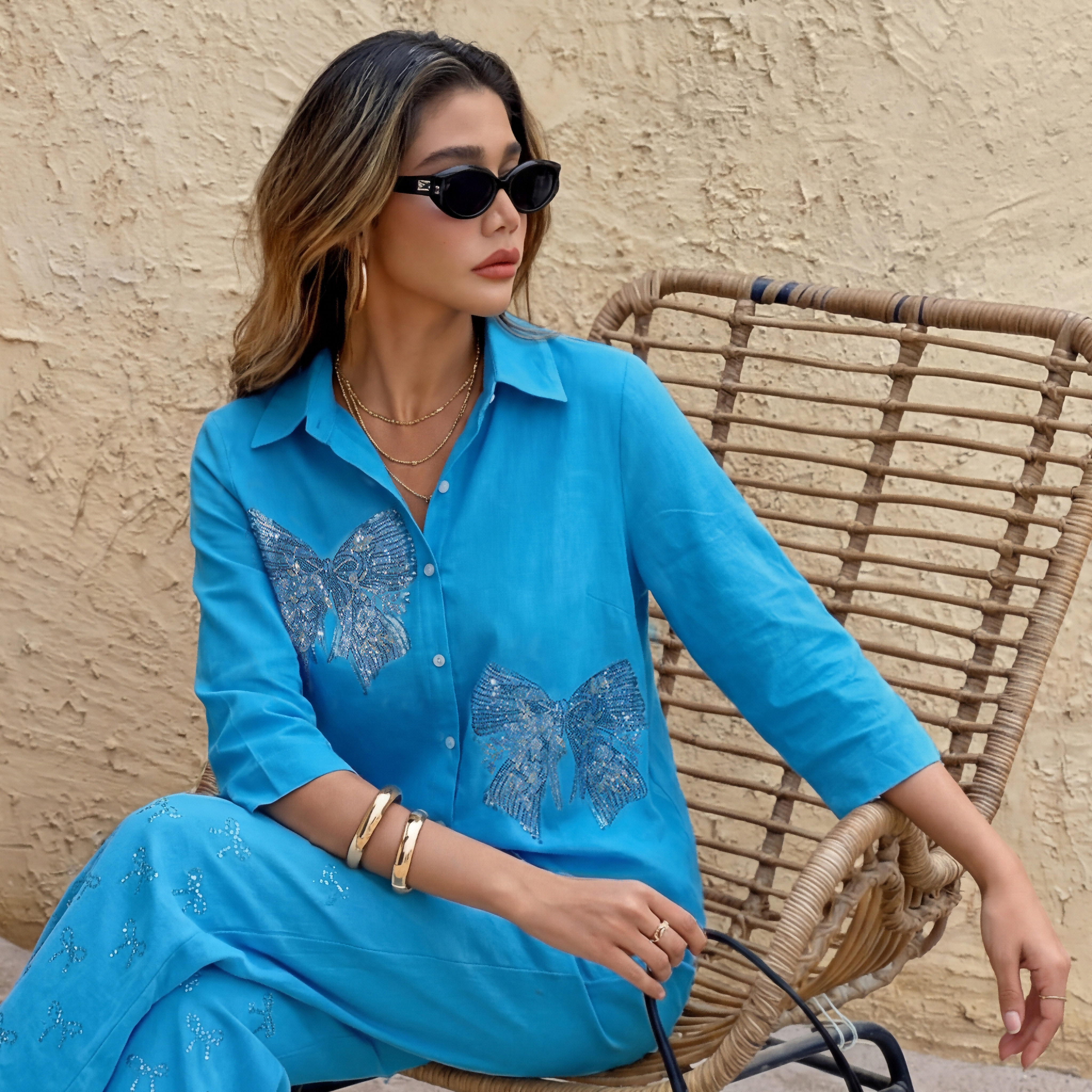 Woman in a blue outfit with butterfly patterns sitting on a wicker chair against a beige wall. TCH Blue Rhinestone Bow Matching Set