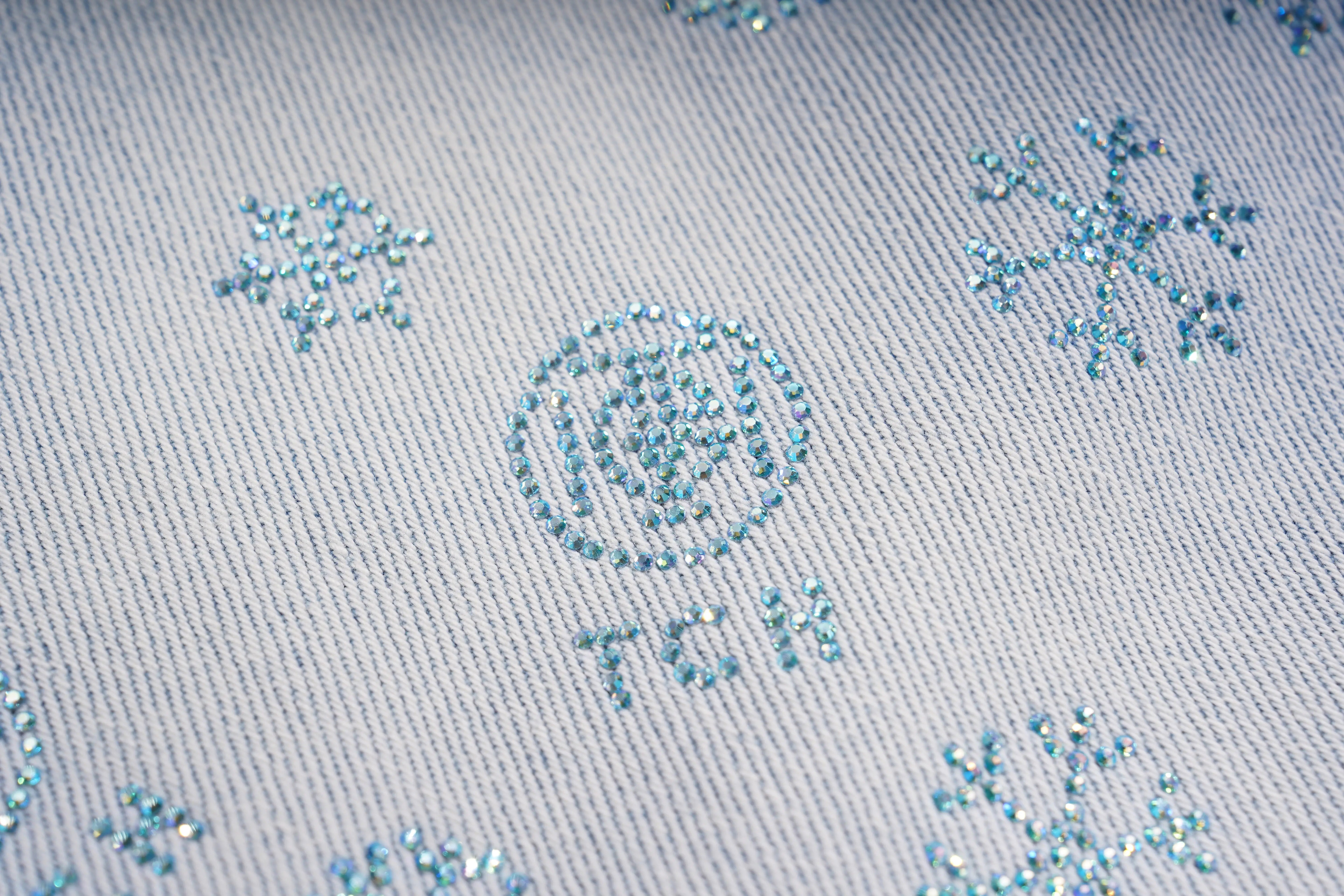 Close-up of a textured fabric surface with decorative elements and 'TCM' branding. TCH Blue Snowflake Rhinestone Denim Jeans