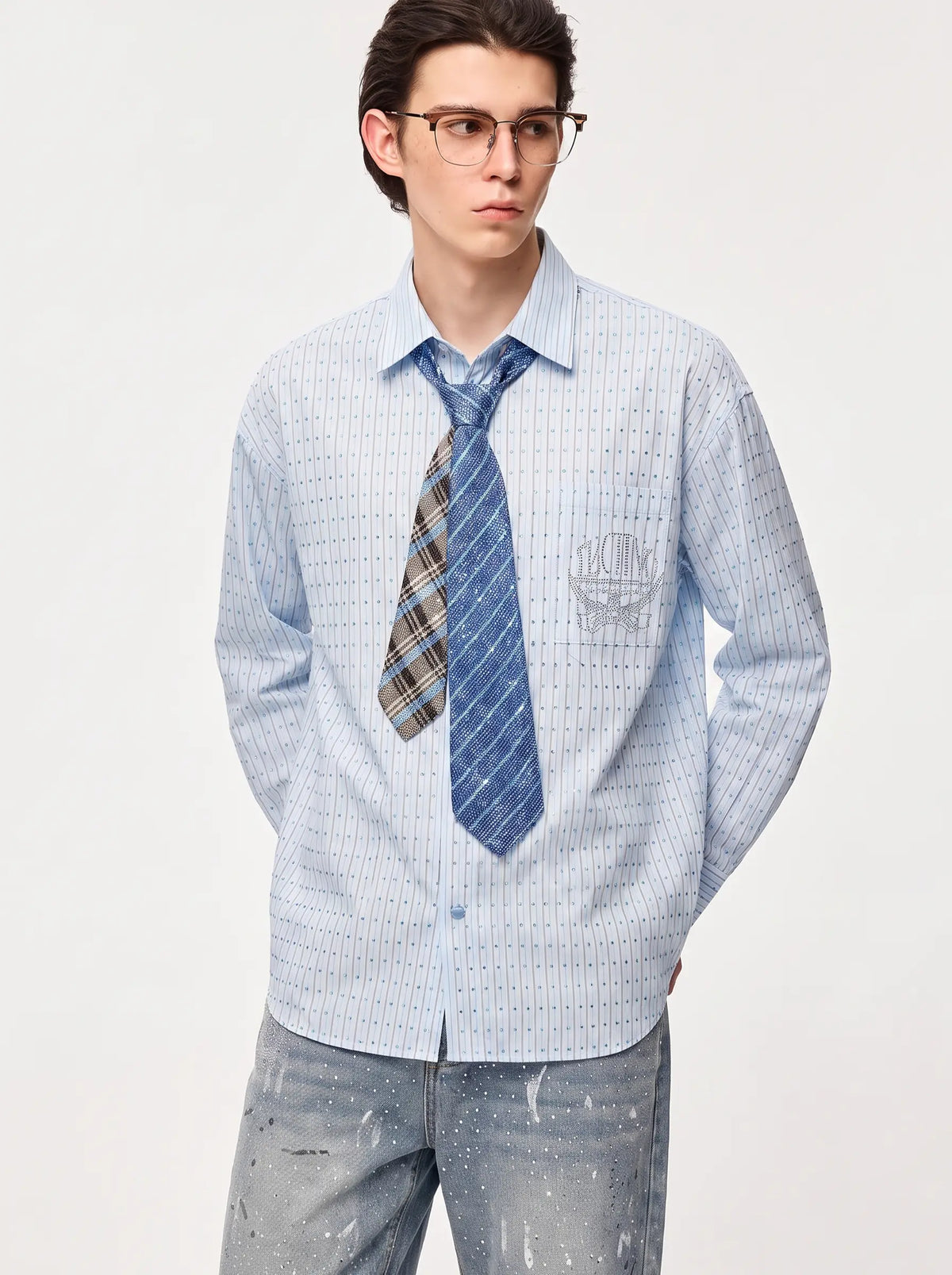 A young man wearing glasses, a light blue and white checkered long-sleeve button-down shirt, a blue patterned tie, and distressed gray jeans, standing against a plain light gray studio background. TCH Blue Striped Tie Detail Button-Up Shirt