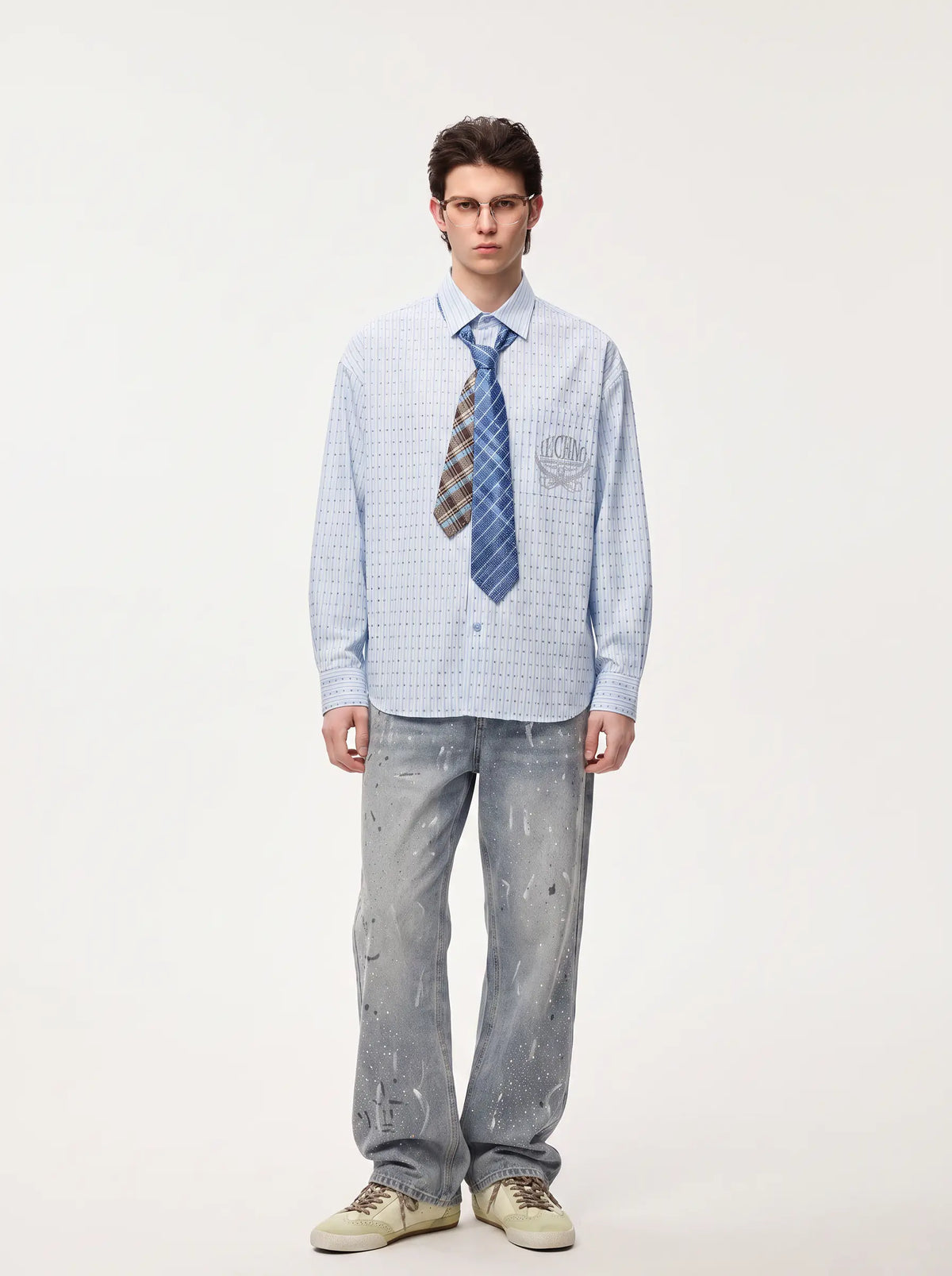 A male model stands against a plain light background wearing a light blue patterned long-sleeve button-up shirt, a blue and brown striped tie, light gray jeans, and beige sneakers. TCH Blue Striped Tie Detail Button-Up Shirt