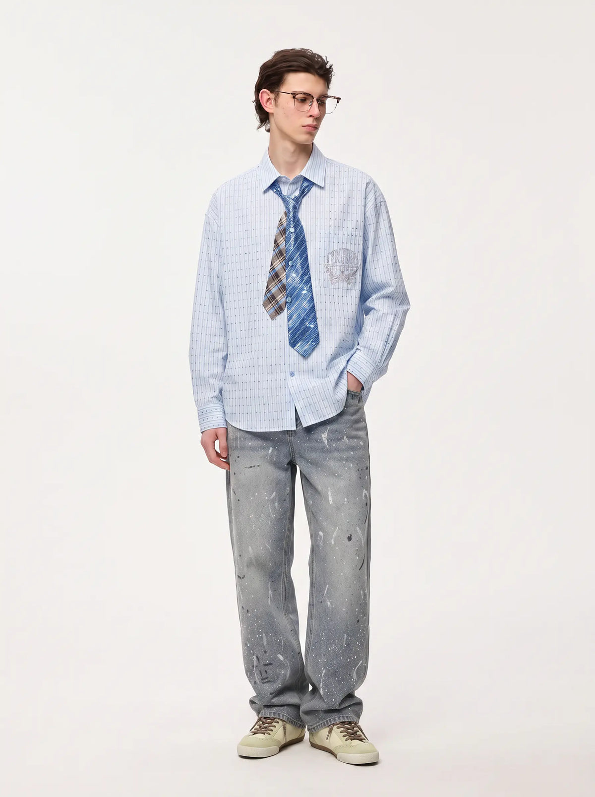 A man modeling a light blue and white checkered long-sleeve button-down shirt, styled with a blue and brown patterned tie, gray paint-splattered jeans, and beige sneakers, against a plain off-white studio background. TCH Blue Striped Tie Detail Button-Up Shirt