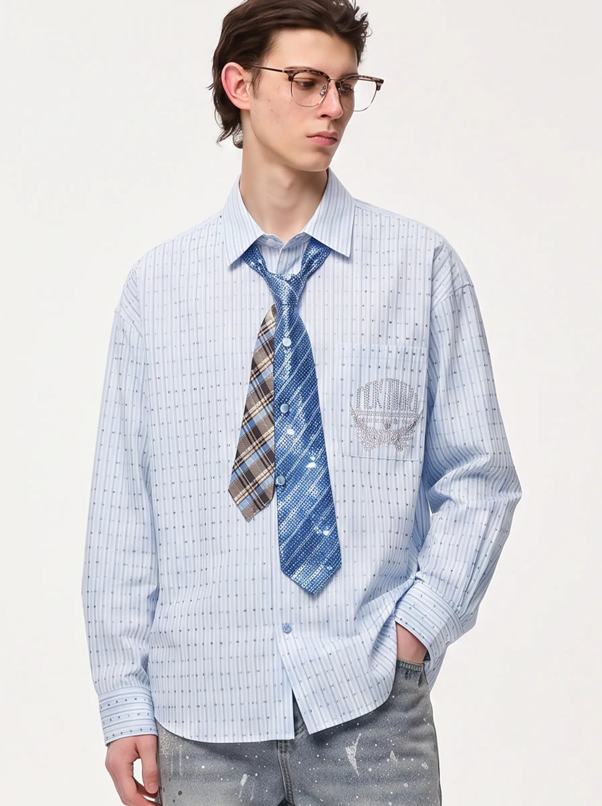 A young man wearing glasses, a light blue and white striped button-down shirt, a blue patterned necktie, and gray jeans, standing against a plain off-white studio background. TCH Blue Striped Tie Detail Button-Up Shirt