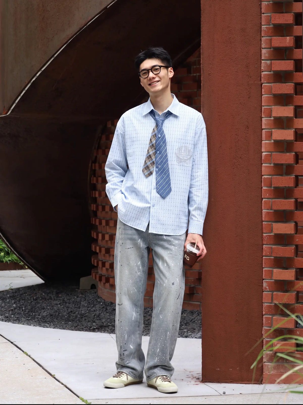 A man wearing a light blue and white checkered long-sleeve shirt, a blue patterned tie, and light gray jeans stands outdoors in front of a brick and metal architectural structure. TCH Blue Striped Tie Detail Button-Up Shirt
