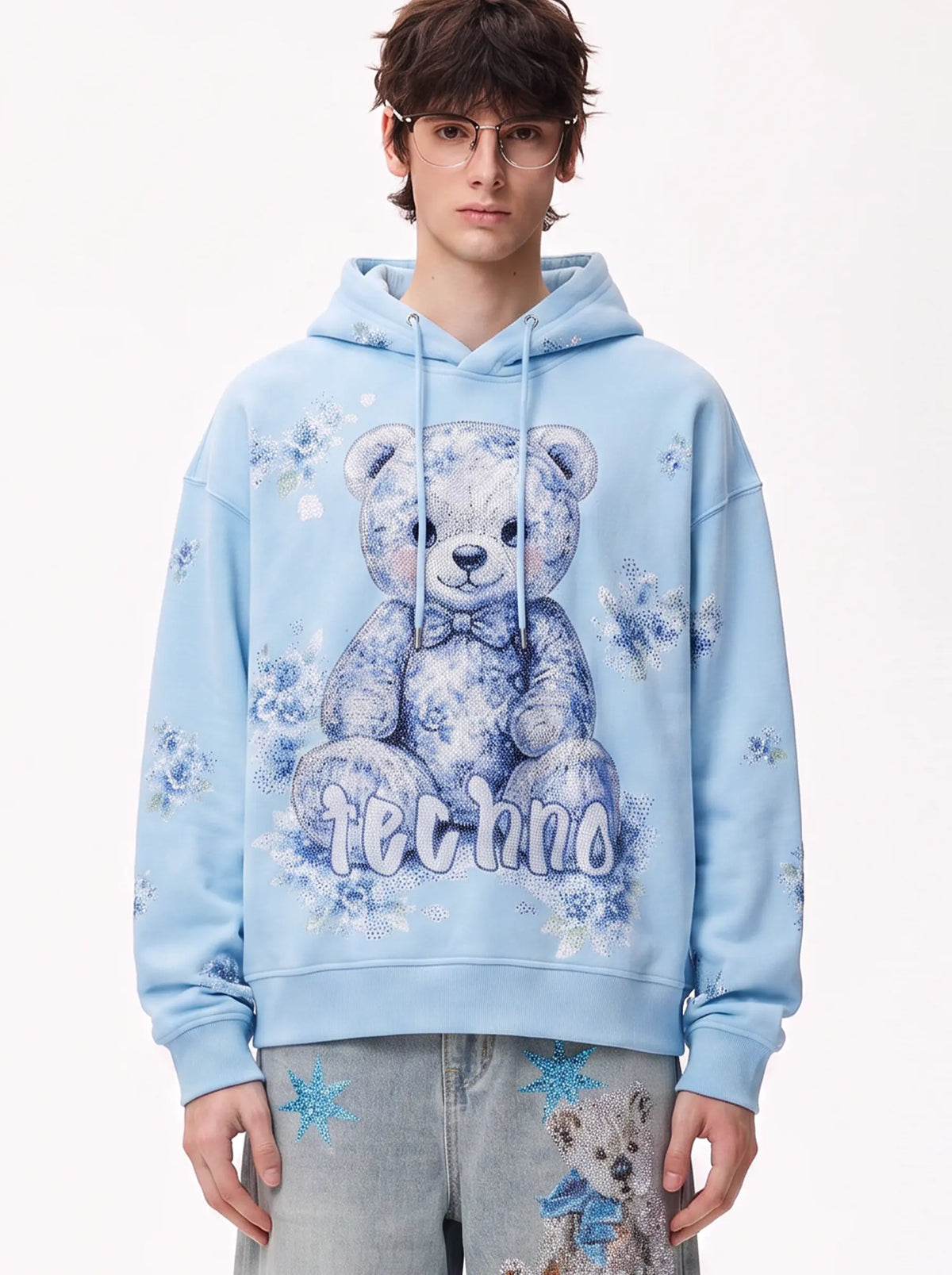 A model wearing a light blue denim jacket with various embroidered patches, including a teddy bear on the left chest and cherries on the sleeve, styled over a white t-shirt and dark pants. TCH Blue Teddy Bear Floral Rhinestone Hoodie