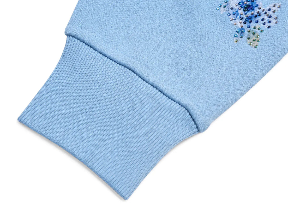 Close-up of a light blue ribbed knit sleeve cuff with a small cluster of blue and silver rhinestones on the upper fabric, shown on a white background. TCH Blue Teddy Bear Floral Rhinestone Hoodie