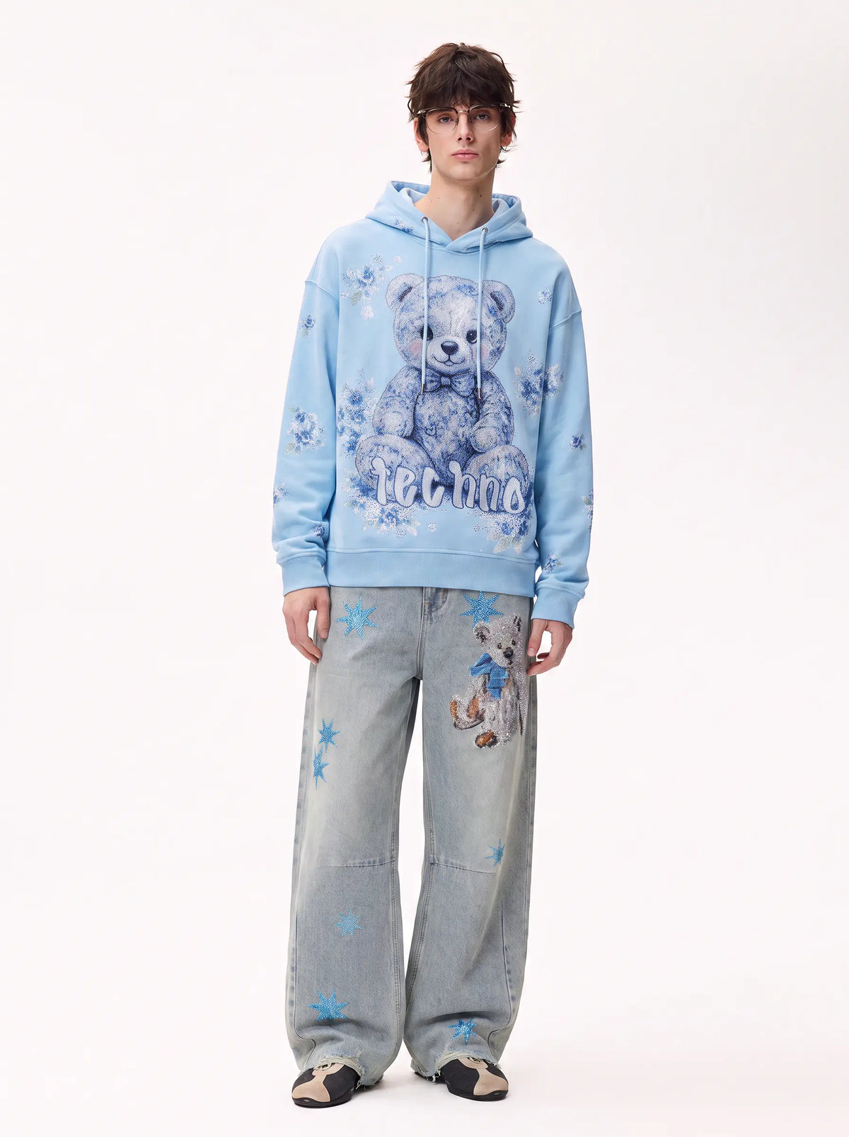 A model stands against a plain white studio background wearing a light blue graphic hoodie with a teddy bear print and the word 'teddy' on the front, paired with wide-leg light blue jeans featuring blue star prints. TCH Blue Teddy Bear Floral Rhinestone Hoodie