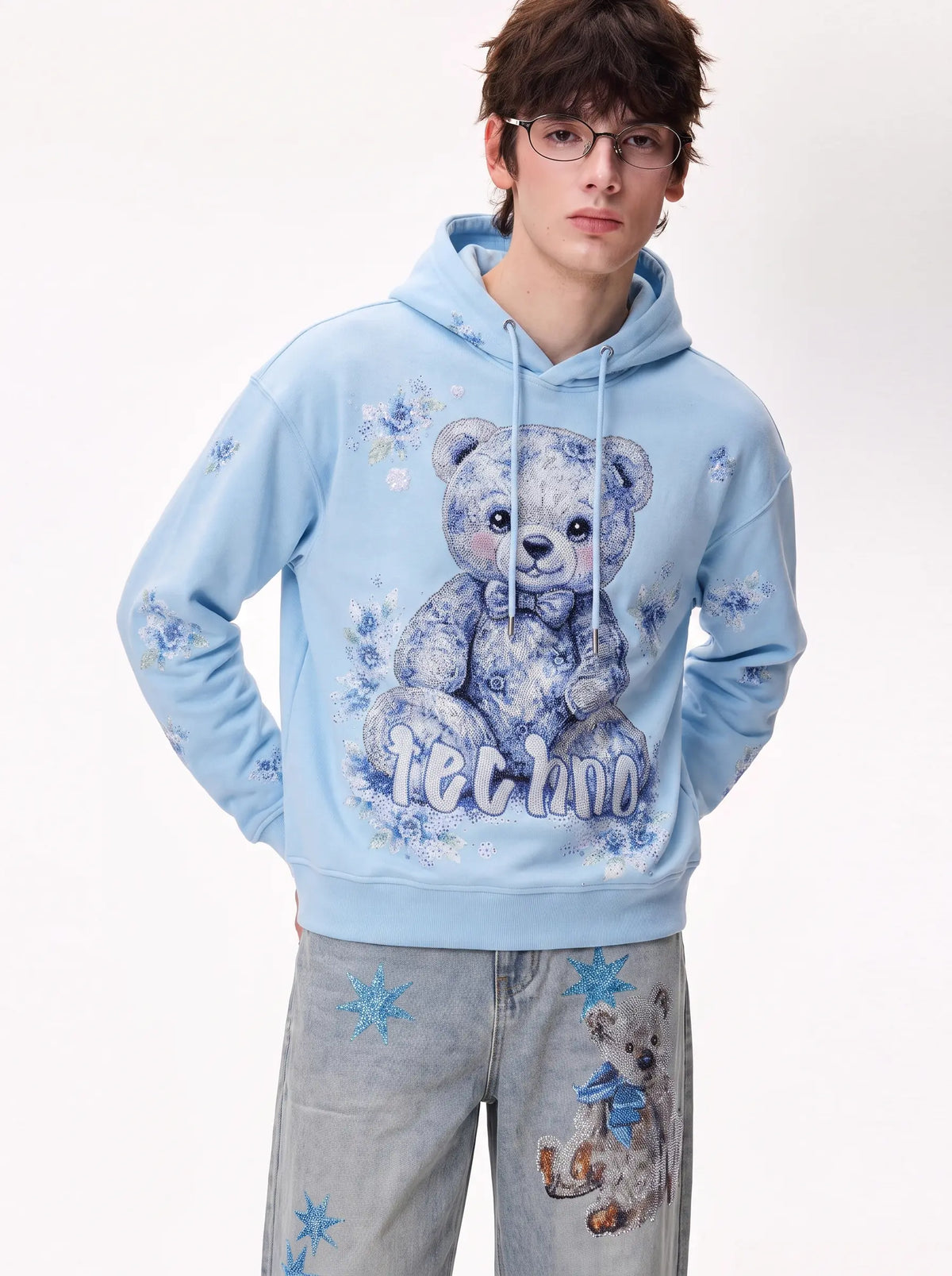 A model wearing a light blue hoodie with a large blue-toned teddy bear graphic and the word 'techno' on the front, paired with light gray jeans featuring blue star and bear prints, against a plain white studio background. TCH Blue Teddy Bear Floral Rhinestone Hoodie