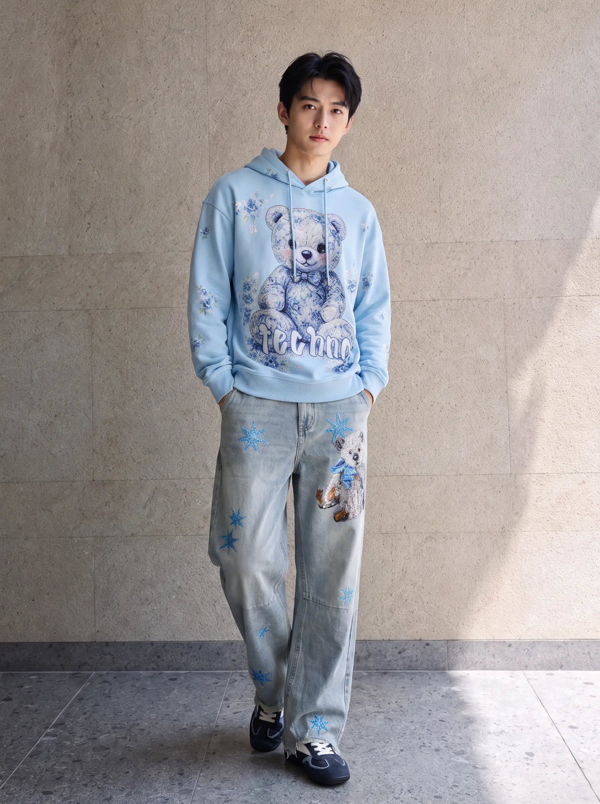 A man wearing a light blue hoodie with a teddy bear graphic and matching light blue jeans with teddy bear prints, standing against a textured beige wall. TCH Blue Teddy Bear Floral Rhinestone Hoodie