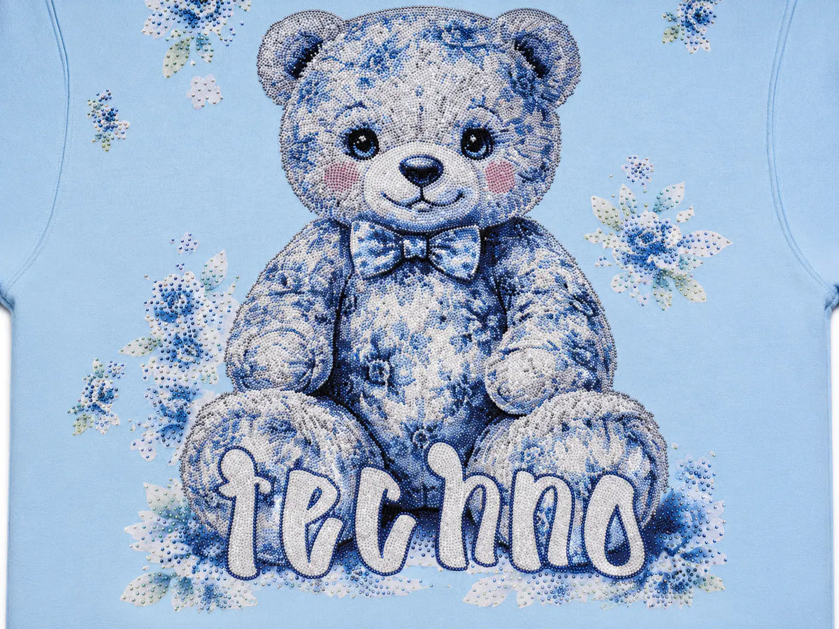 A close-up of a light blue t-shirt featuring a large graphic of a teddy bear with a blue and white floral pattern, a matching bow tie, and the word 'techno' written in stylized white letters across the bottom. TCH Blue Teddy Bear Floral Rhinestone Hoodie