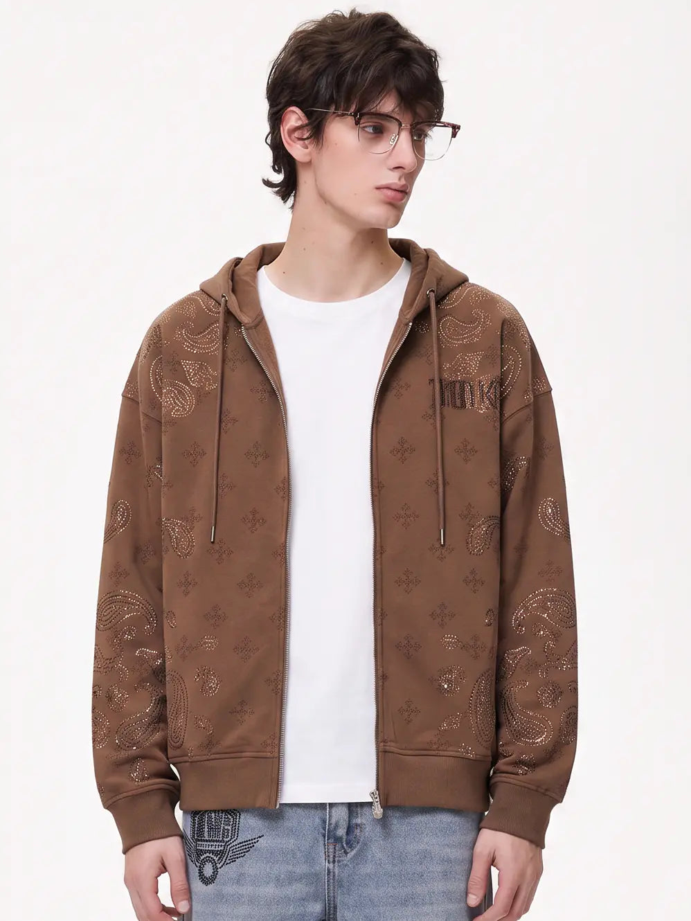A model wearing a light blue denim jacket with various embroidered patches, including a teddy bear on the left chest and cherries on the sleeve, styled over a white t-shirt and dark pants. TCH Brown Rhinestone Paisley Zip Up Hoodie