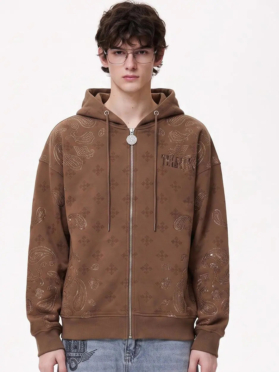 A young man wearing a brown zip-up hoodie with an all-over tonal paisley and monogram pattern, paired with light blue jeans, against a plain white studio background. TCH Brown Rhinestone Paisley Zip Up Hoodie