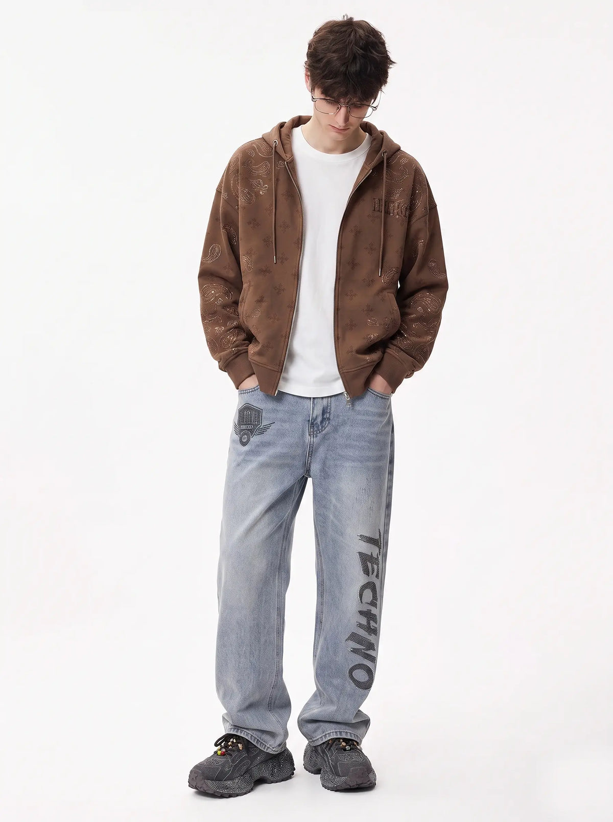 A man wearing a brown hooded zip-up jacket over a white t-shirt, light-wash blue jeans with 'TECHNO' printed down one leg, and dark gray sneakers, standing against a plain white studio background. TCH Brown Rhinestone Paisley Zip Up Hoodie