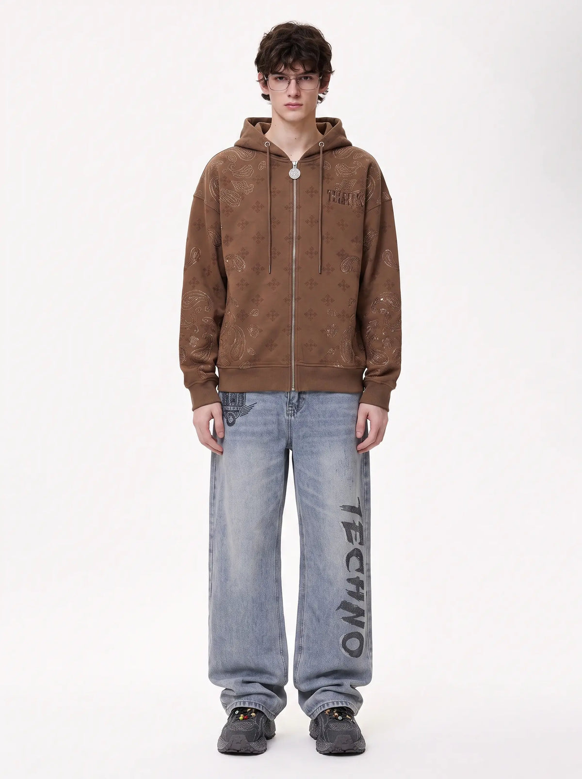 A model stands against a plain white studio background wearing a brown hooded sweatshirt with a distressed, mottled texture and light-wash blue jeans featuring vertical black 'TECHNO' text on the leg. TCH Brown Rhinestone Paisley Zip Up Hoodie