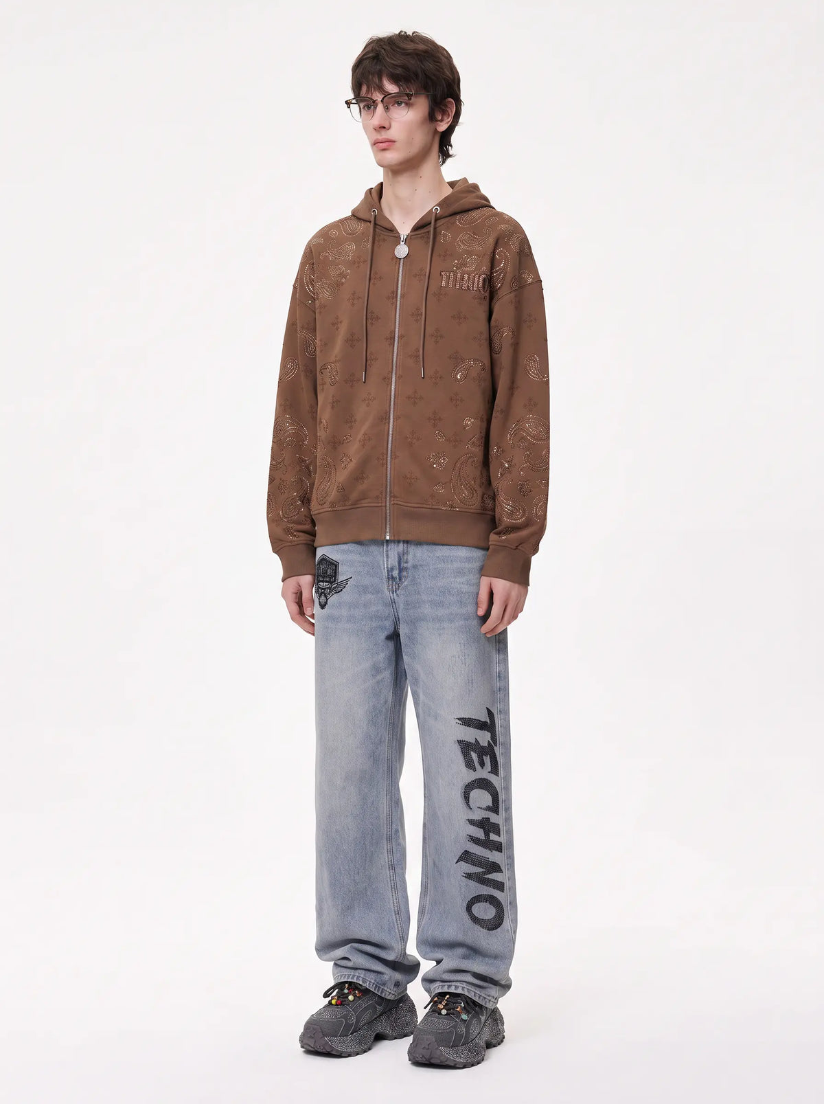 A model stands against a plain white studio background wearing a brown zip-up hoodie with a distressed, faded texture, light-wash blue jeans with 'TECHNO' printed vertically on the leg, and dark gray sneakers.