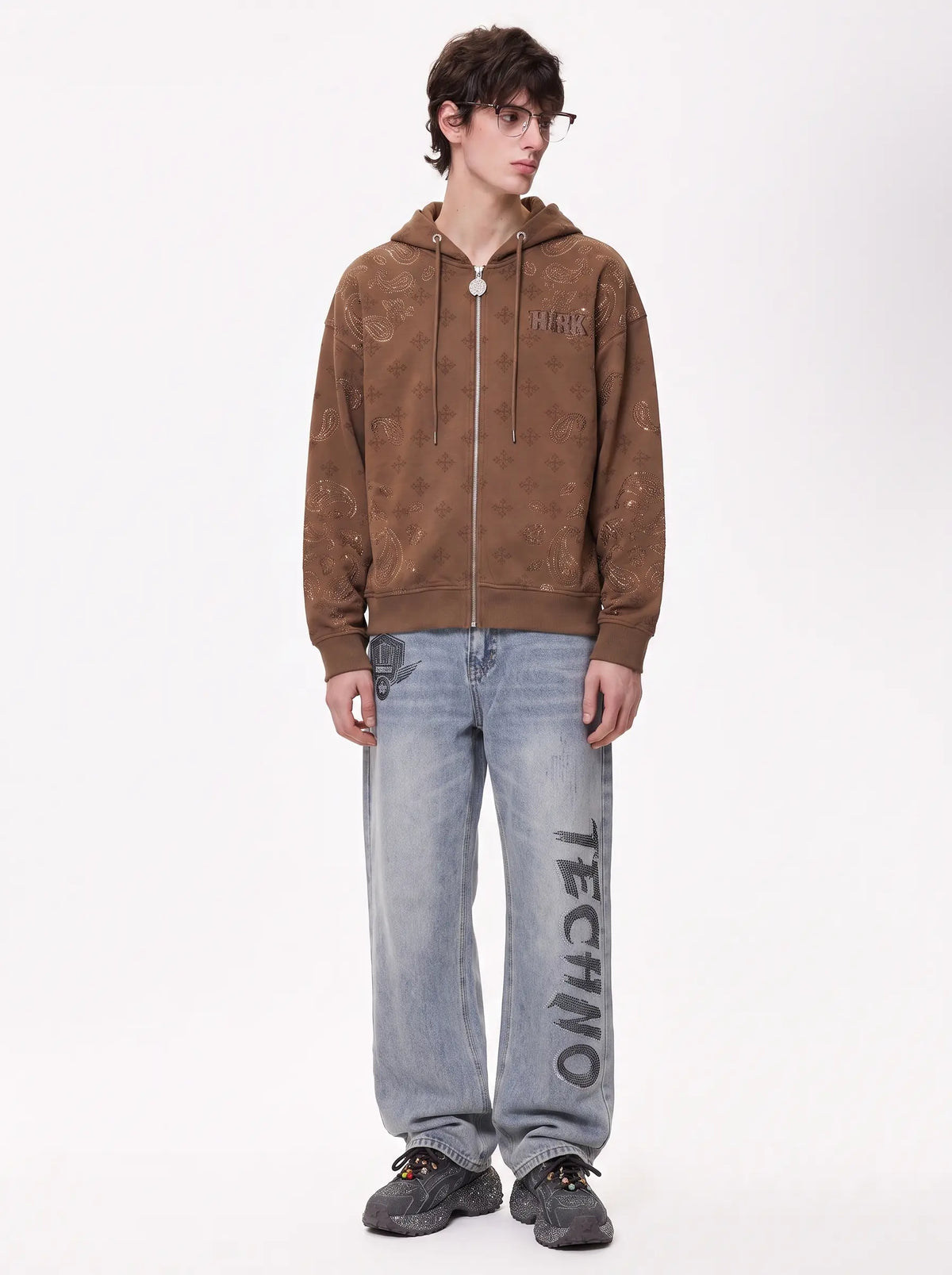 A model stands against a plain white studio background wearing a brown zip-up hoodie with a distressed texture, light-wash blue jeans with 'TECHNO' printed down the leg, and dark sneakers. TCH Brown Rhinestone Paisley Zip Up Hoodie