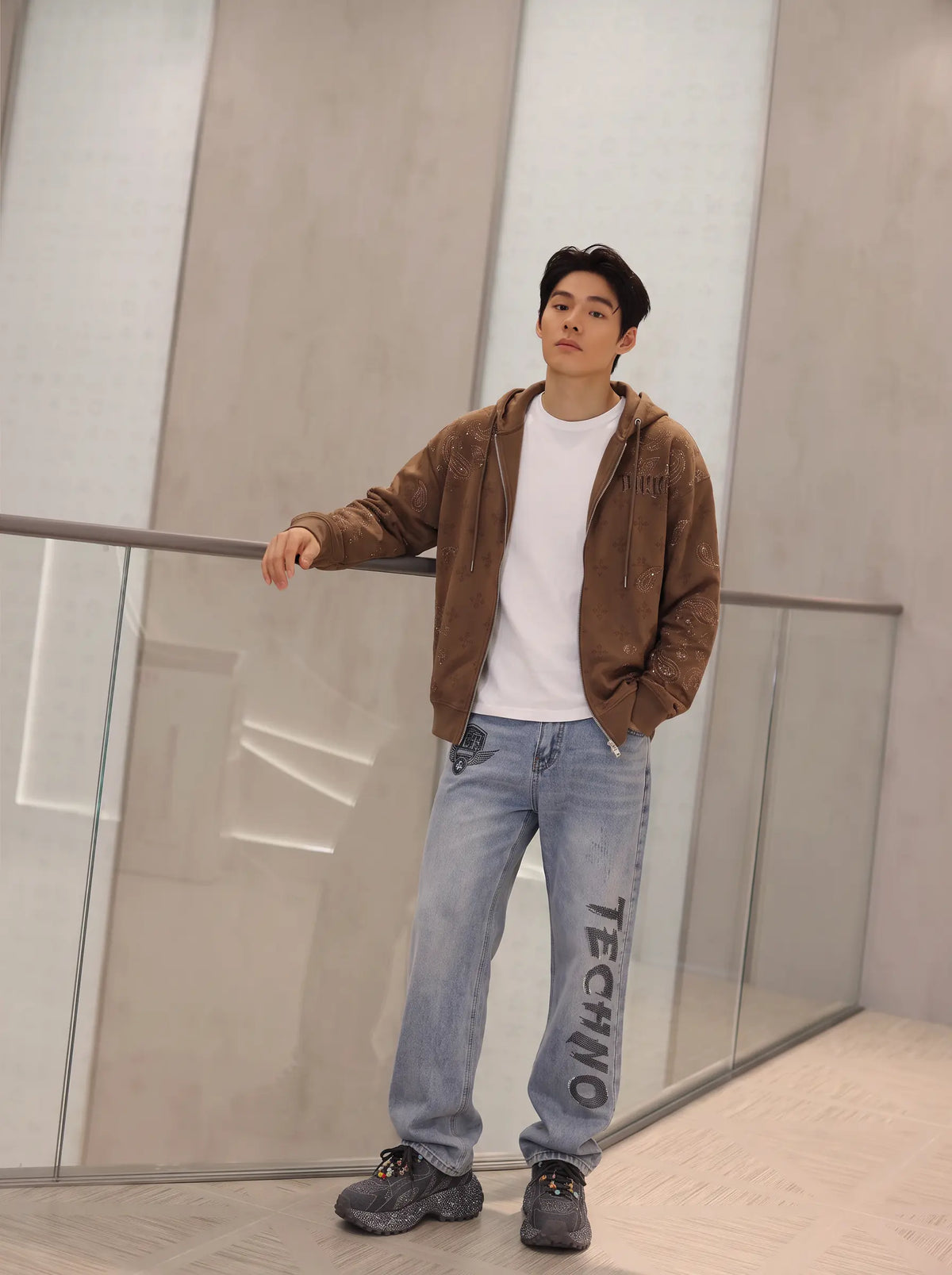 A man wearing a brown zip-up hoodie over a white t-shirt, light blue jeans with 'TECHNO' printed on the leg, and dark sneakers, leaning on a glass railing in a modern indoor setting. TCH Brown Rhinestone Paisley Zip Up Hoodie