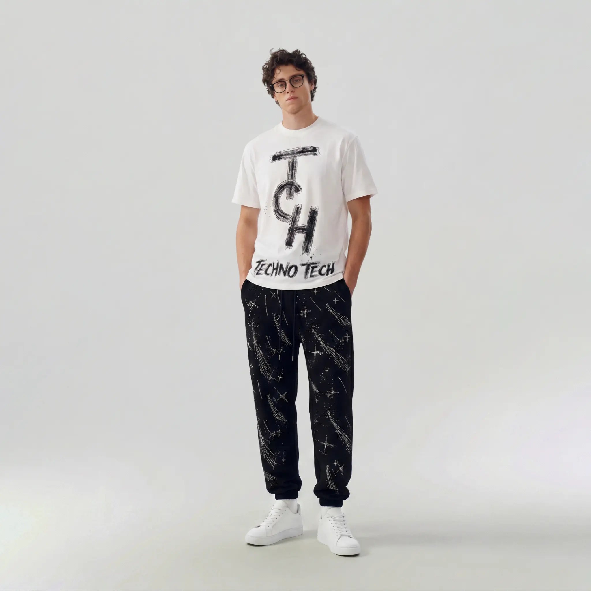 A man modeling a white short-sleeve graphic t-shirt with a black 'Tech' logo and 'ZECNO TECH' text, paired with black pants featuring a white starry pattern and white sneakers, against a light gray studio background. TCH Brush Logo Graphic Oversized T-Shirt