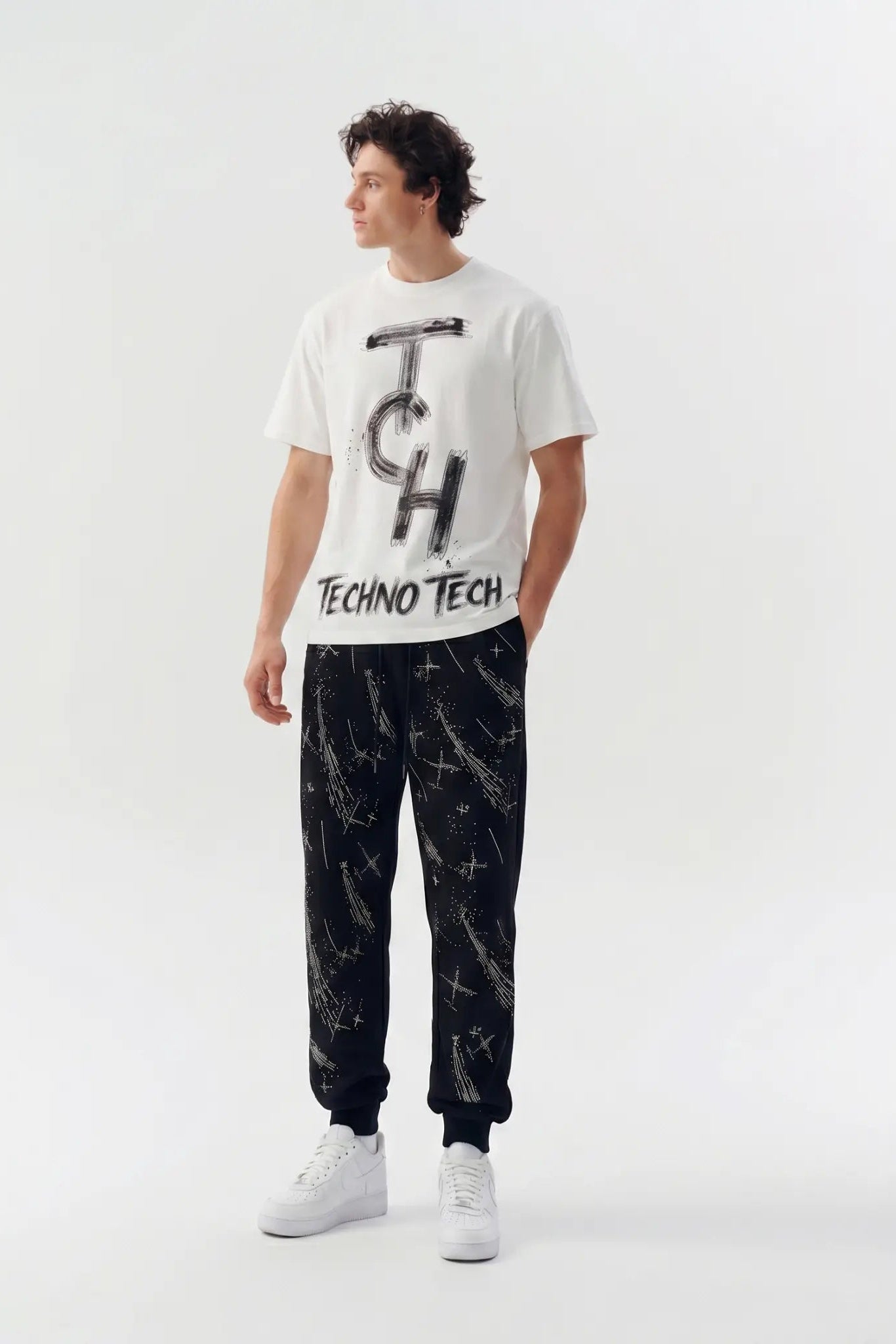 A man modeling a white Techno Tech t-shirt with a black graphic print and black pants featuring a white abstract pattern, standing against a light gray studio background. TCH Brush Logo Graphic Oversized T-Shirt