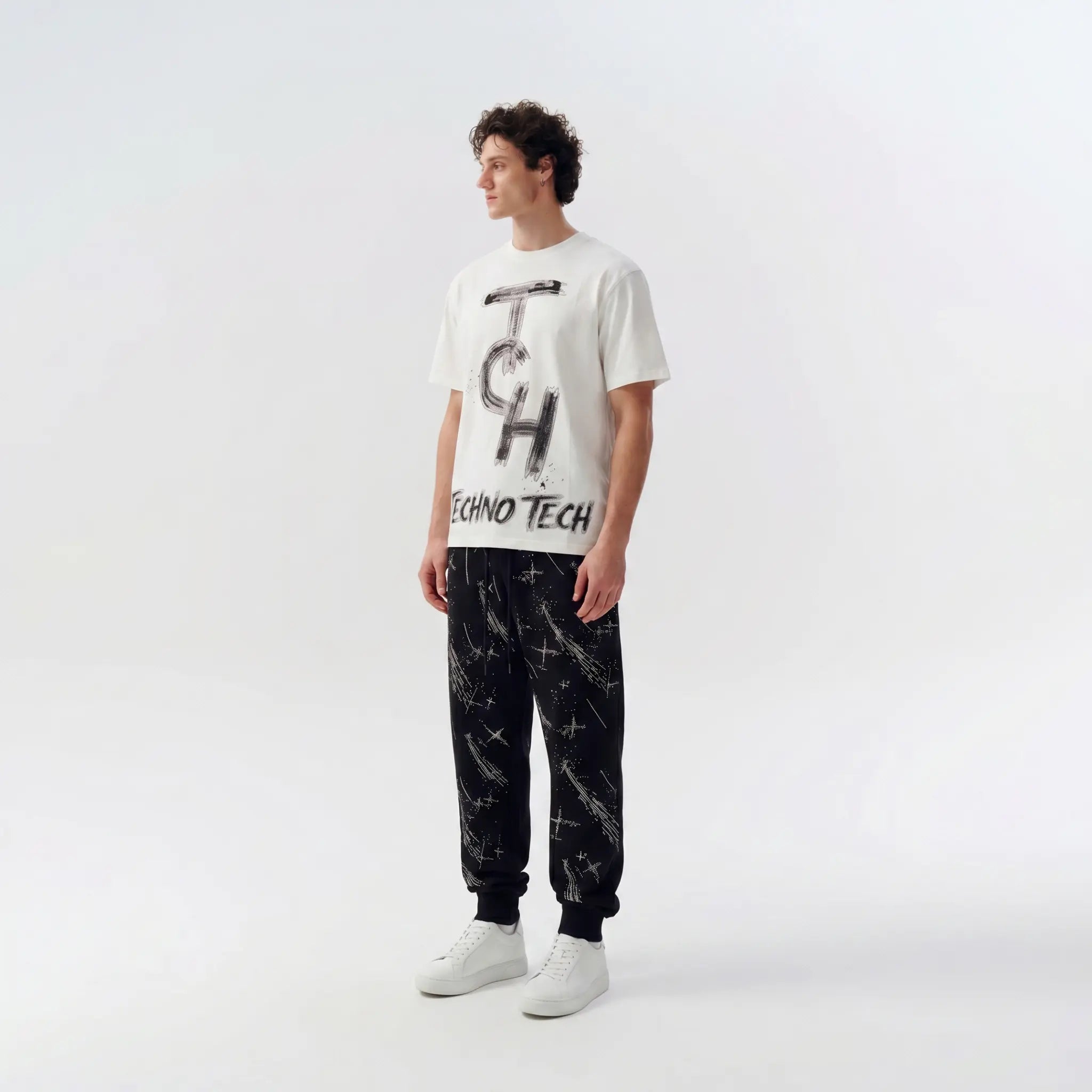 A male model stands in a studio wearing a white graphic T-shirt with black 'TECH' lettering, black jogger pants with a white paint-splatter pattern, and white sneakers against a light gray background. TCH Brush Logo Graphic Oversized T-Shirt
