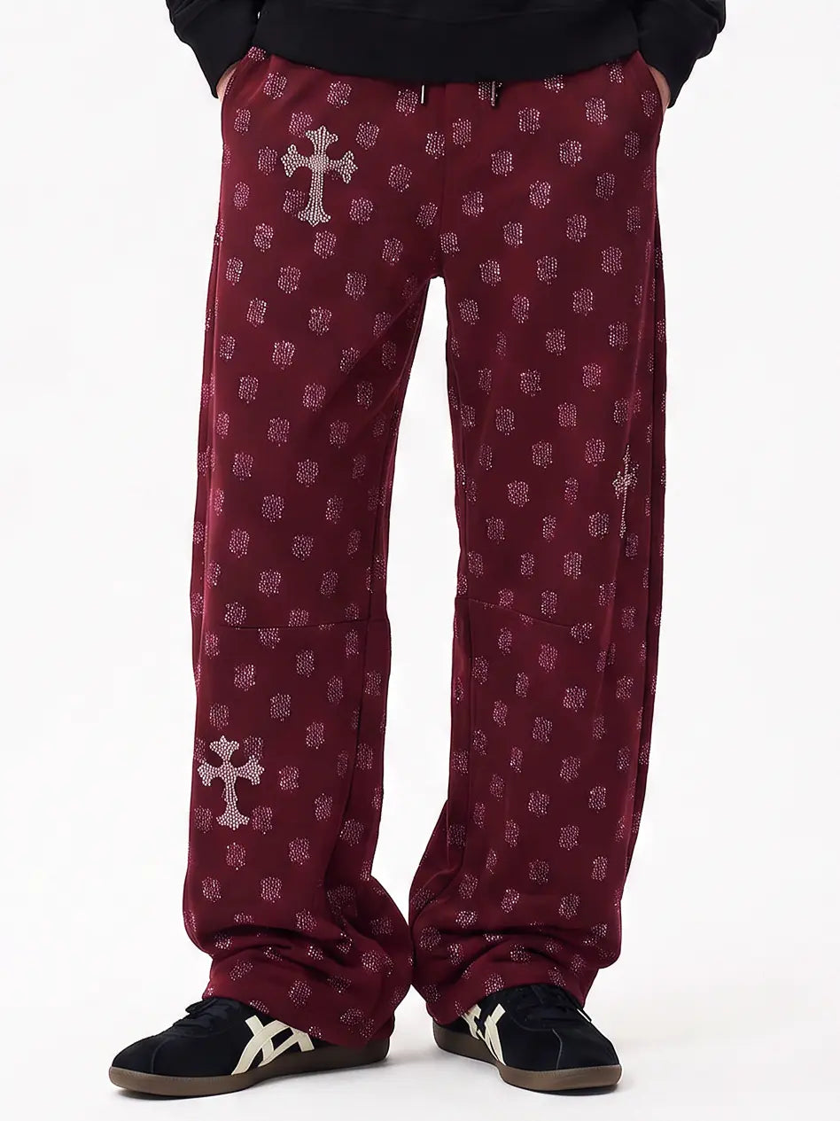 A person wearing burgundy pants with a repeating small cross pattern and larger silver cross motifs on the thighs, paired with black sneakers against a plain white studio background. TCH Burgundy Paisley Print Wide-Leg Sweatpants with Cross Graphic