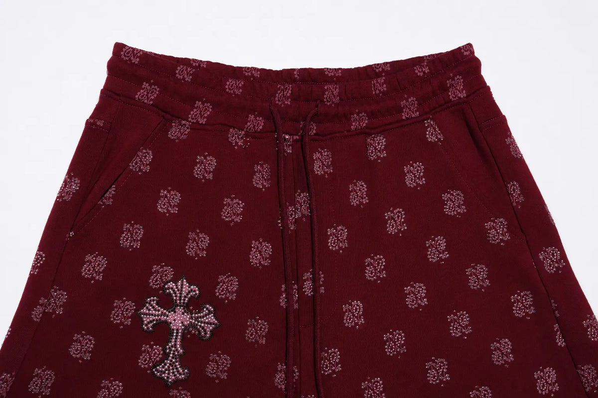 A close-up of burgundy drawstring pants featuring a repeating tonal floral pattern and a large embellished cross motif on the left leg, set against a plain white background. TCH Burgundy Paisley Print Wide-Leg Sweatpants with Cross Graphic