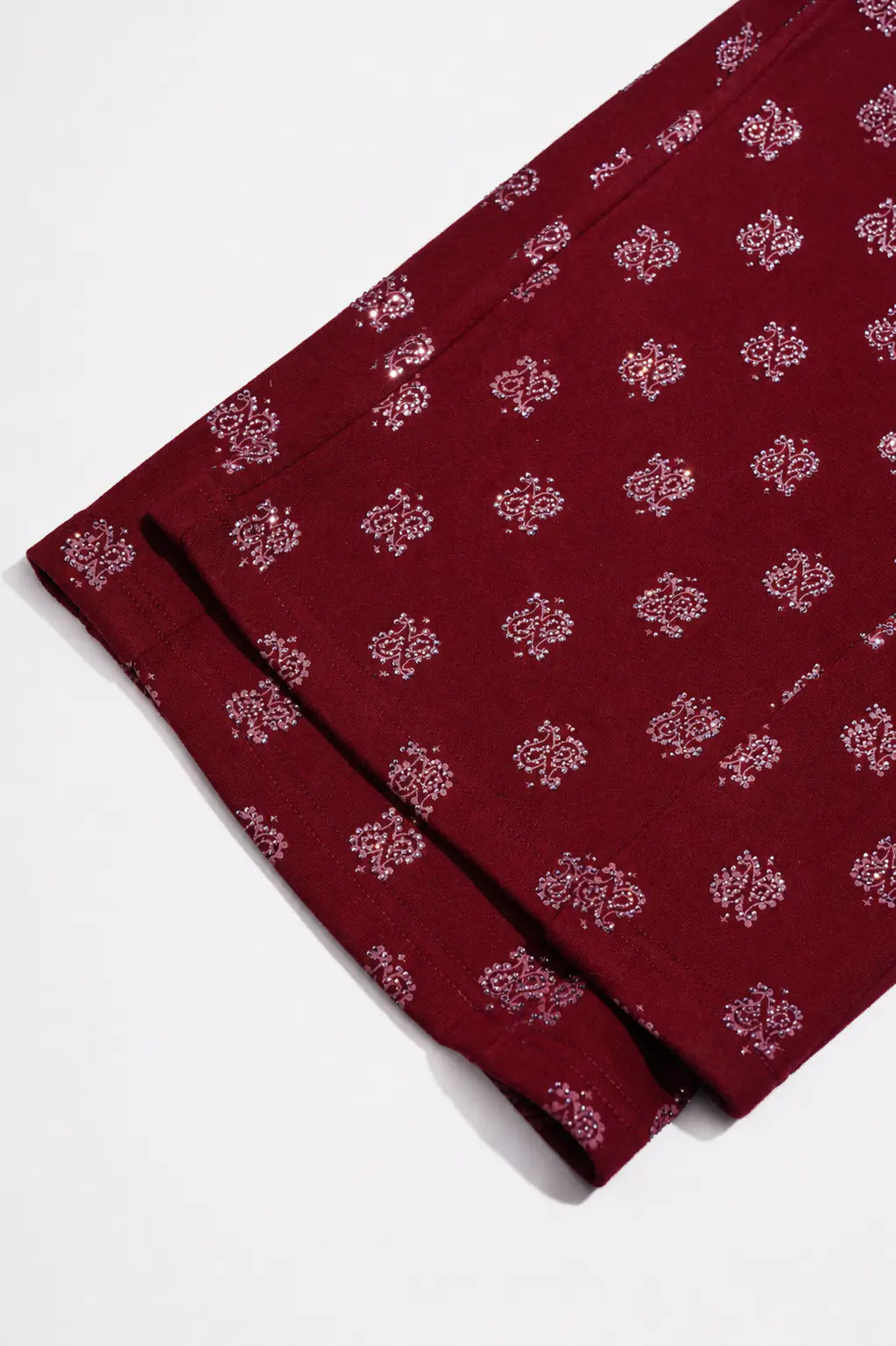 A close-up of a folded deep maroon fabric with a repeating light pink floral block print, shown on a clean white background. TCH Burgundy Paisley Print Wide-Leg Sweatpants with Cross Graphic