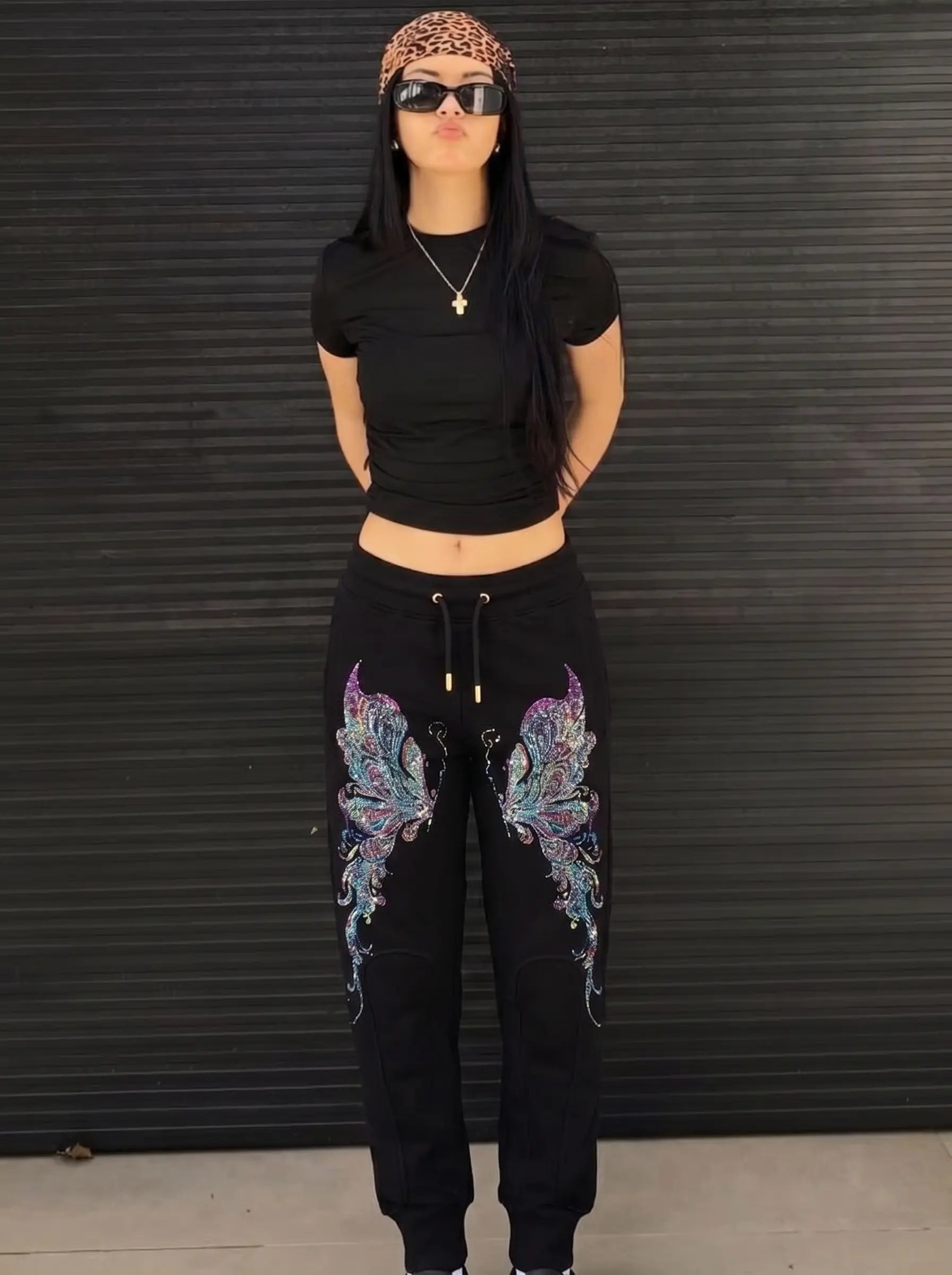 A woman stands in front of a dark corrugated metal wall wearing a black short-sleeve crop top, black sweatpants with large multicolor butterfly graphics on the thighs, a leopard-print headband, and black sunglasses. TCH Butterfly Rhinestone Graphic Black Jogger Pants