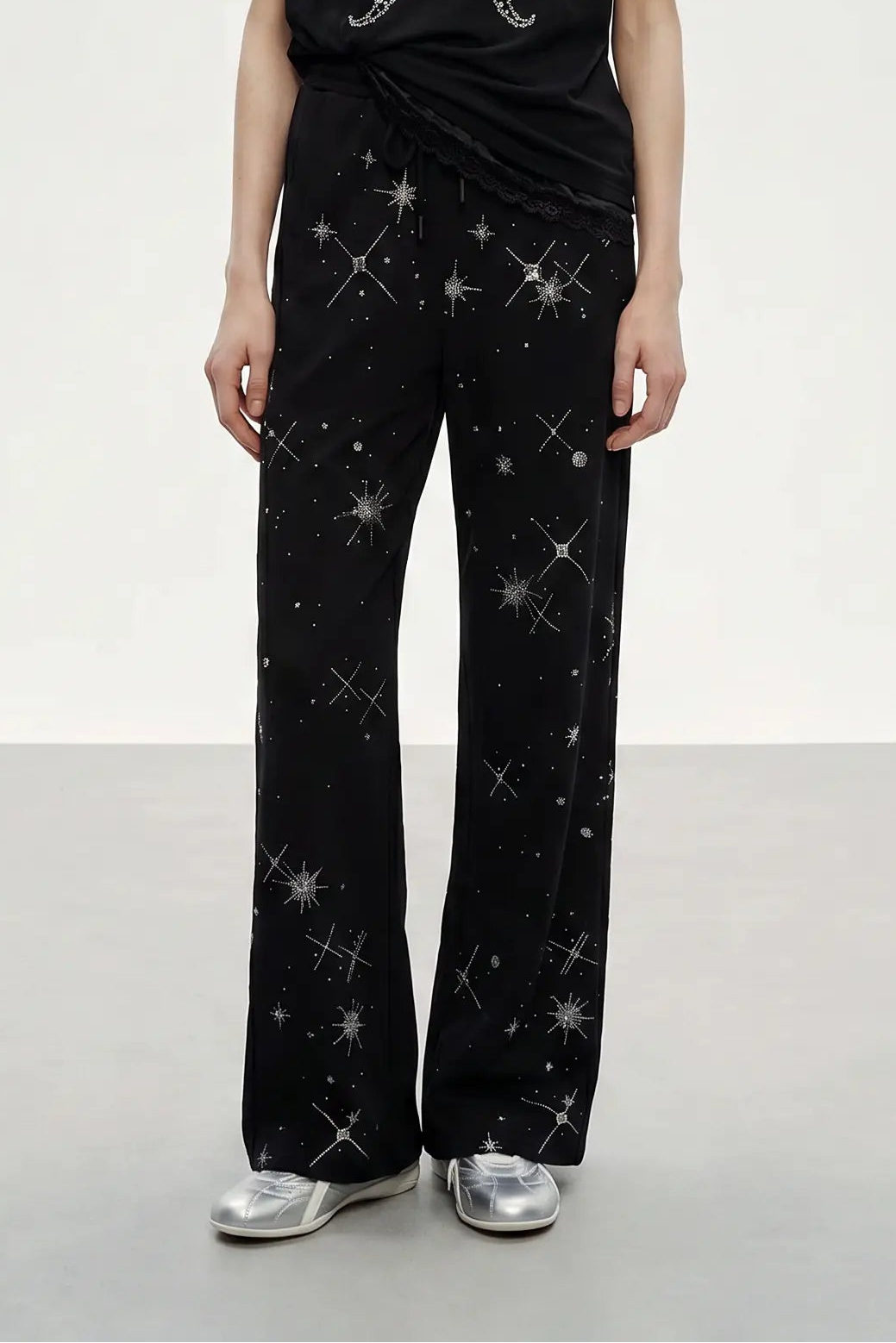 A model is shown from the waist down wearing black wide-leg pants with a silver starry constellation print, paired with a black top and silver sneakers, against a neutral studio background. TCH Celestial Rhinestone Wide-Leg Sweatpants