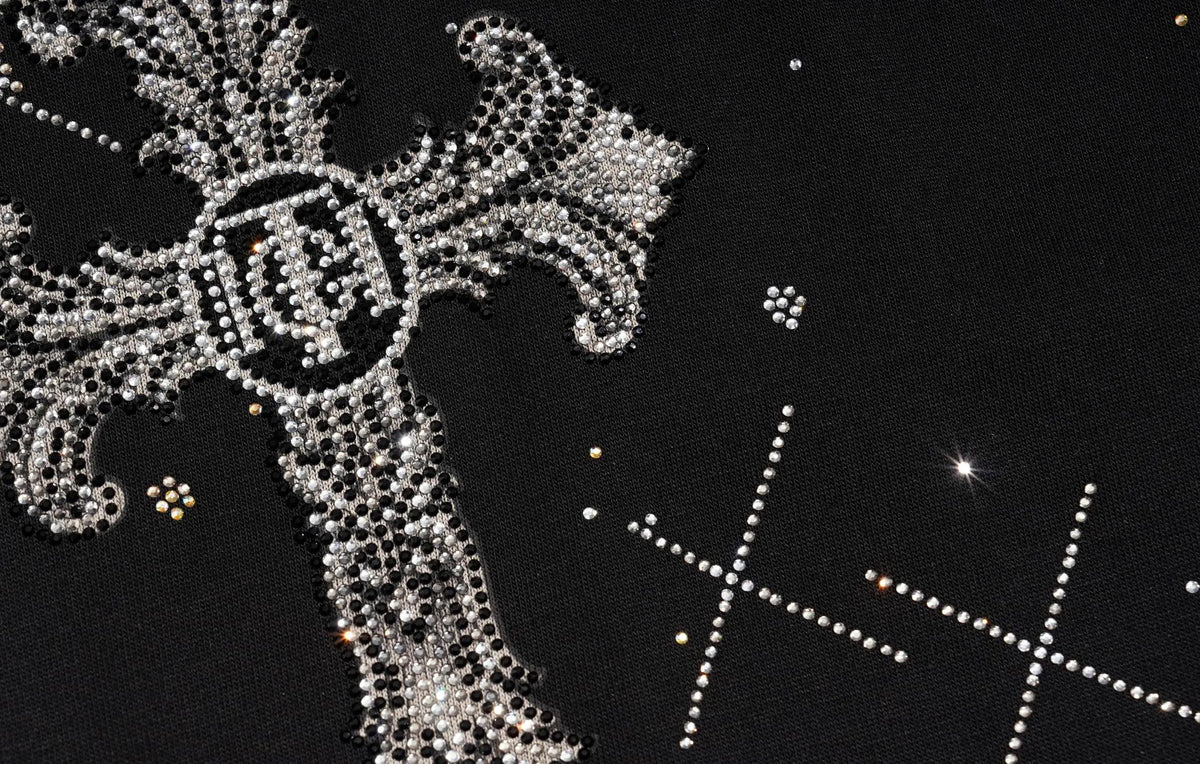 A close-up of a black garment featuring a large ornate silver rhinestone cross with a circular emblem in the center, plus smaller rhinestone star and cross motifs on the fabric. TCH Celestial Rhinestone Wide-Leg Sweatpants