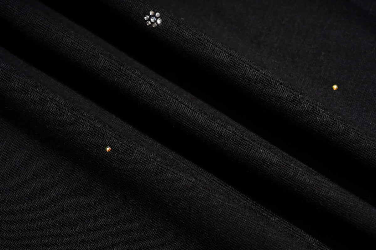 Close-up of a black textured fabric with a small silver floral embellishment and tiny gold dots, draped in soft folds. TCH Celestial Rhinestone Wide-Leg Sweatpants